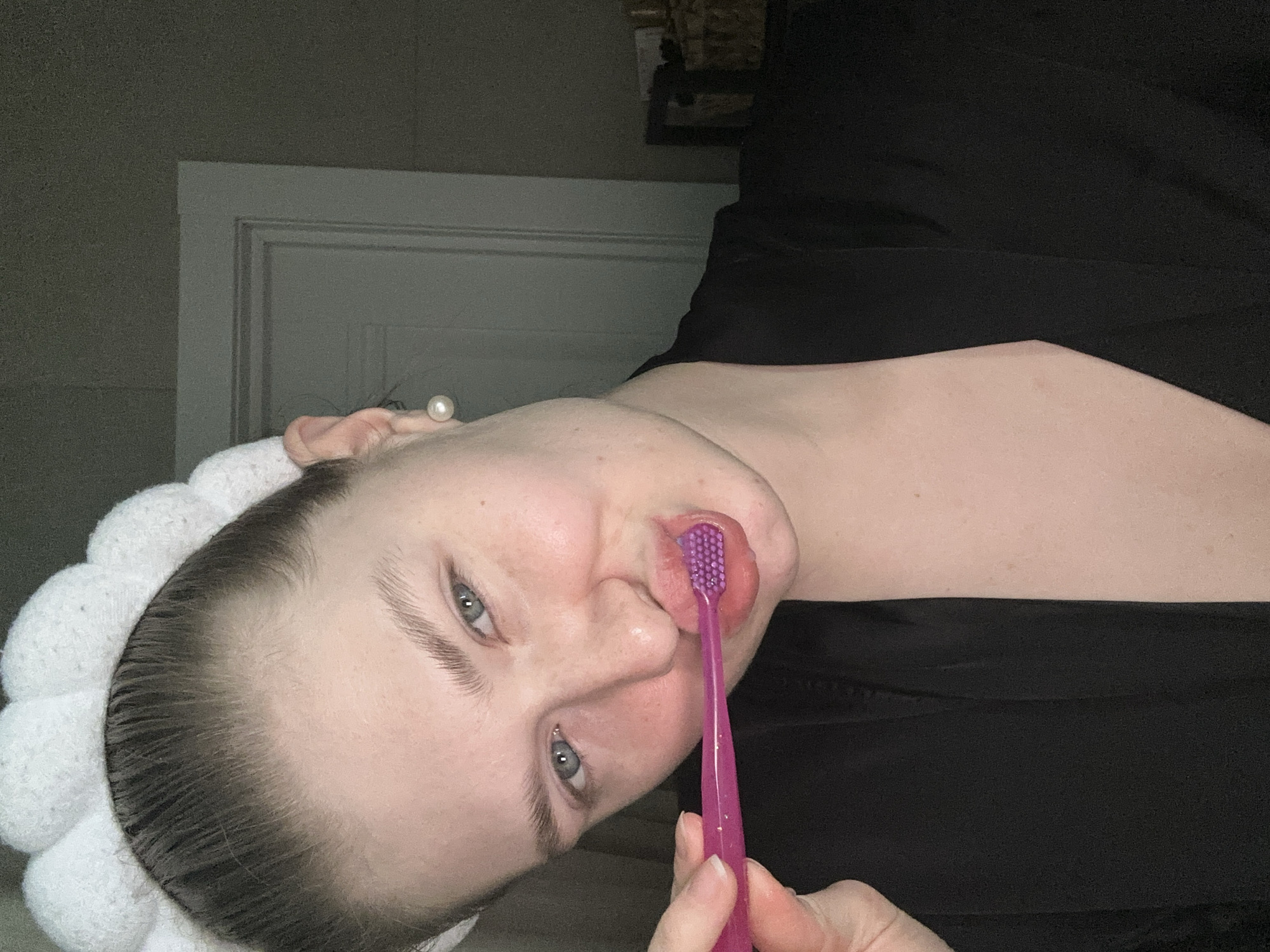 A young adult female is depicted in an indoor setting within Kazan, Russia, facing the camera from the chest up. She wears a white, plush, scalloped headband, a black robe, and a pearl stud earring in her visible right ear. Her light brown hair is pulled back. She holds a translucent pink toothbrush with purple bristles against her lower lip with her right hand, which has neatly manicured fingernails. Her blue eyes maintain direct contact with the viewer, and her lips form a slight smirk. The background on the right side features a light grey door frame against a darker grey wall. Below the door frame, a dark, indistinct piece of furniture, possibly a shelf, holds a light-colored woven object. The scene is dimly lit.