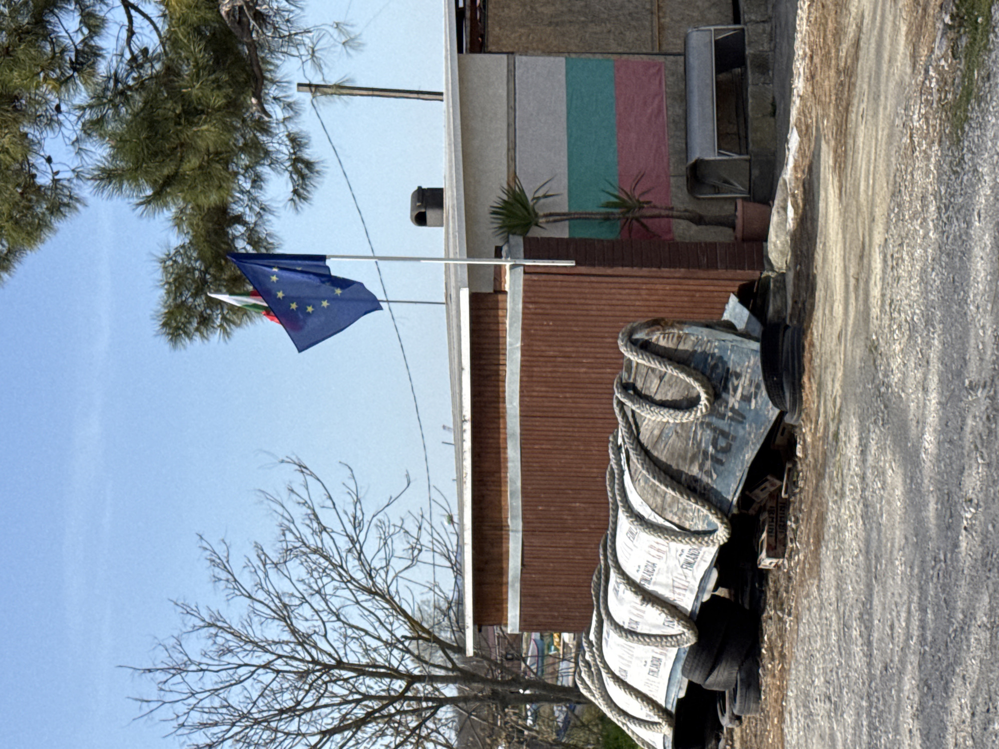 This media file captures an outdoor scene, likely in Varna, Bulgaria, under a bright, clear blue sky. The image is oriented vertically (rotated 90 degrees clockwise from original view).
In the upper left, lush green foliage of a pine tree fills part of the frame. Below the tree, two flags fly on a flagpole. The European Union flag, blue with a circle of twelve yellow stars, is prominently displayed, with the Bulgarian flag (white, green, and red horizontal stripes) partially visible beneath it.
The central background features a building with different textured sections. An upper part appears to be made of light-colored panels. Below this, a section of the wall is decorated with three distinct vertical panels in light grey, teal, and reddish-pink. A small potted palm tree stands in front of these colored panels. Further to the right, another building section shows a brown, possibly corrugated or slatted, surface.
In the mid-ground and foreground, a dark, boat-like object or large container is covered by a white tarp or canvas, around which a thick rope is coiled. The canvas repeatedly displays the text "FINLANDIA." Below this object, several tires are visible. Behind it, wooden crates are stacked, one of which clearly shows the text "MINI."
The right side and foreground of the image are dominated by a gravel path or unpaved road. The scene suggests a daytime setting, with no human activity or specific events taking place.