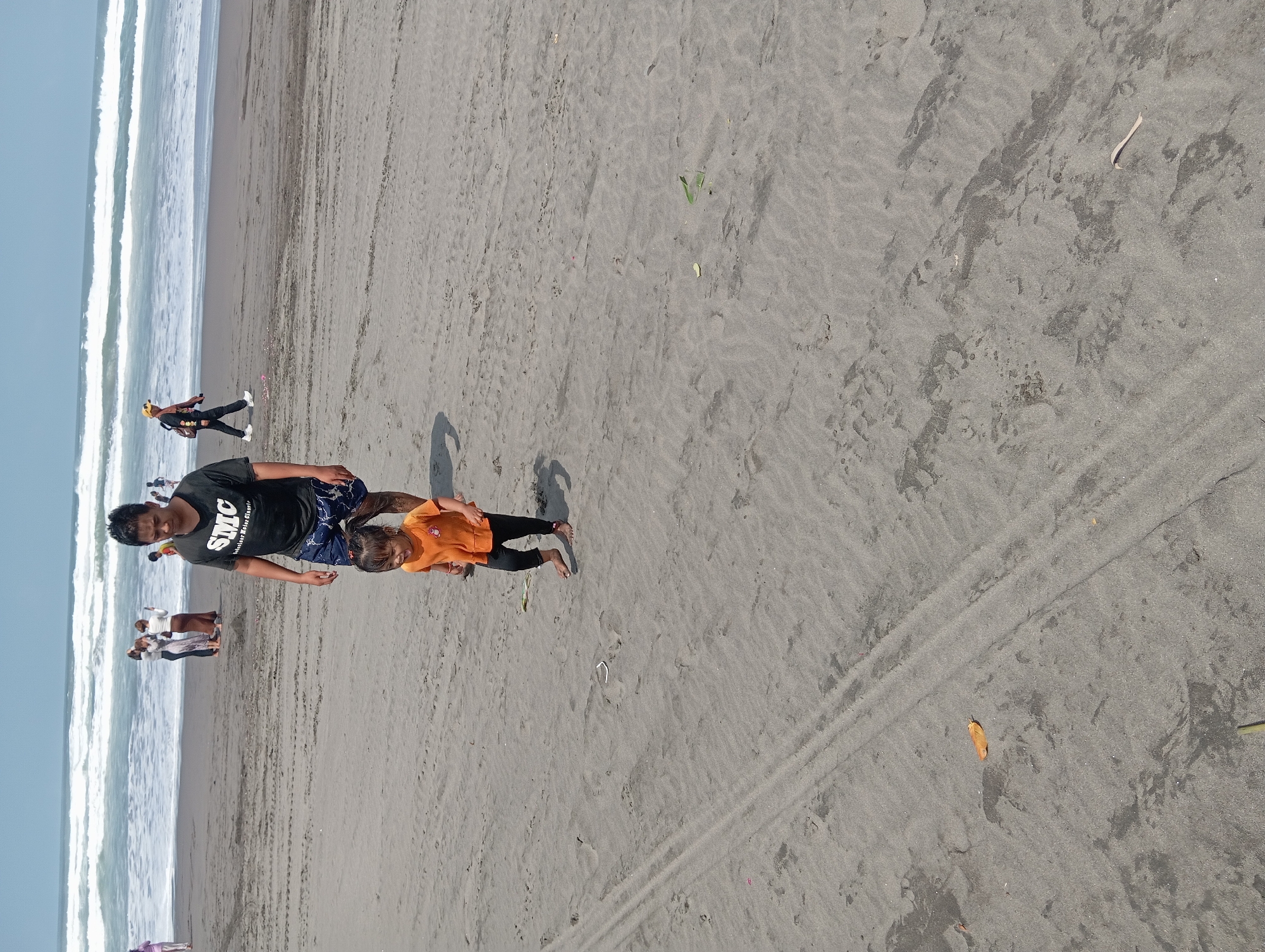 The image shows a beach scene at Parangtritis, Indonesia. In the foreground, a man in a black t-shirt with "SMC" logo and patterned shorts walks on the dark sand, with a young child in an orange shirt and black pants running slightly ahead of him. The child's tongue is sticking out. In the background, the ocean with white waves stretches to the horizon under a clear sky. Several other people are visible further down the beach, some standing and talking, and one person walking away from the camera. The sand has tire tracks imprinted on it, suggesting vehicle access to the beach. The overall scene is of a casual outing at the beach.