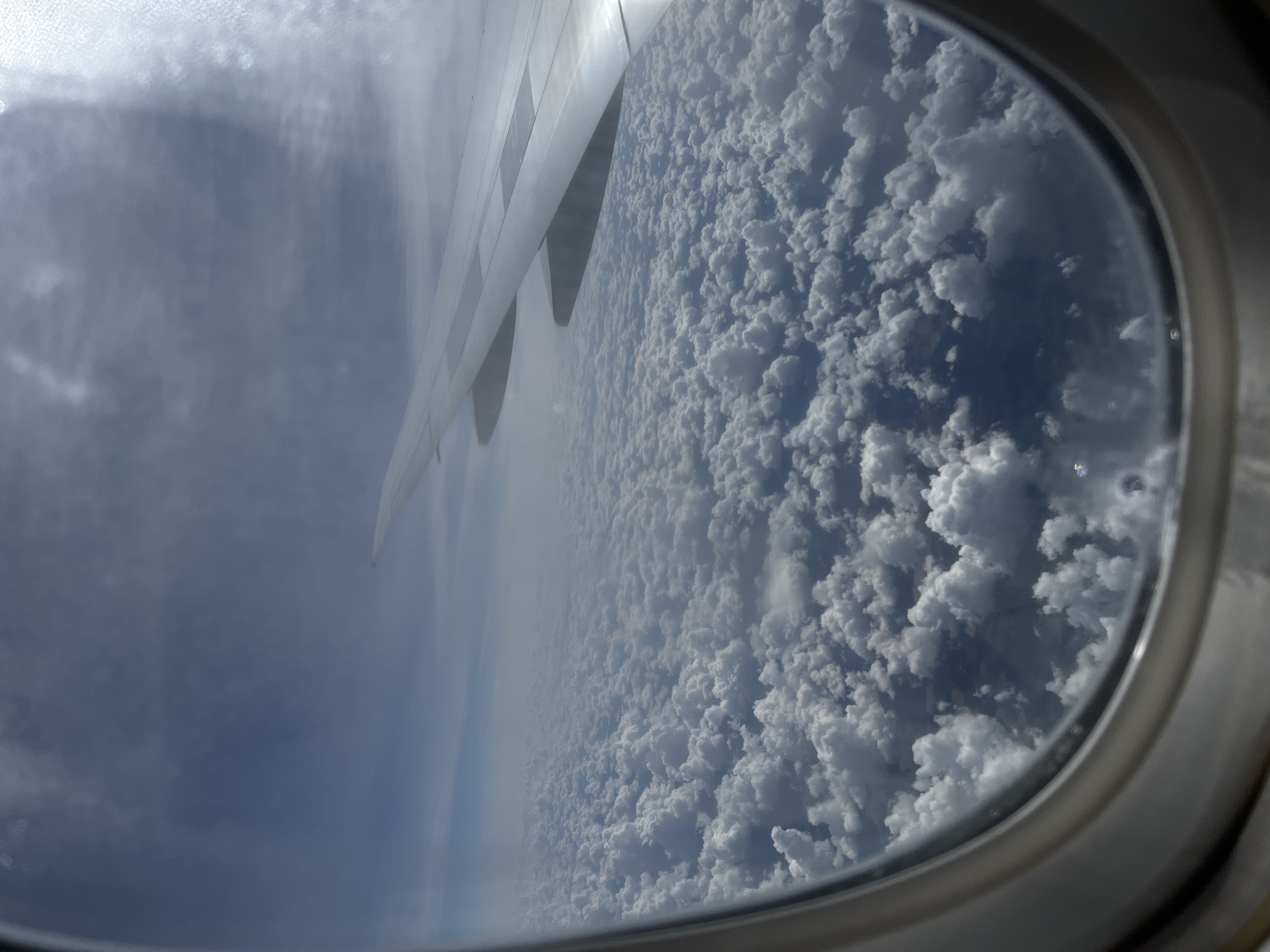 An aerial view of fluffy white clouds is seen through an airplane window. The right side of an airplane wing is visible, extending into the frame from the top right corner. The sky above the clouds is a pale blue, with some hazy clouds present. The image is taken from an elevated perspective, looking down upon a vast expanse of clouds. The location is specified as Sukhanikha, Russia, though no specific landmarks or city details are discernible from the image itself.