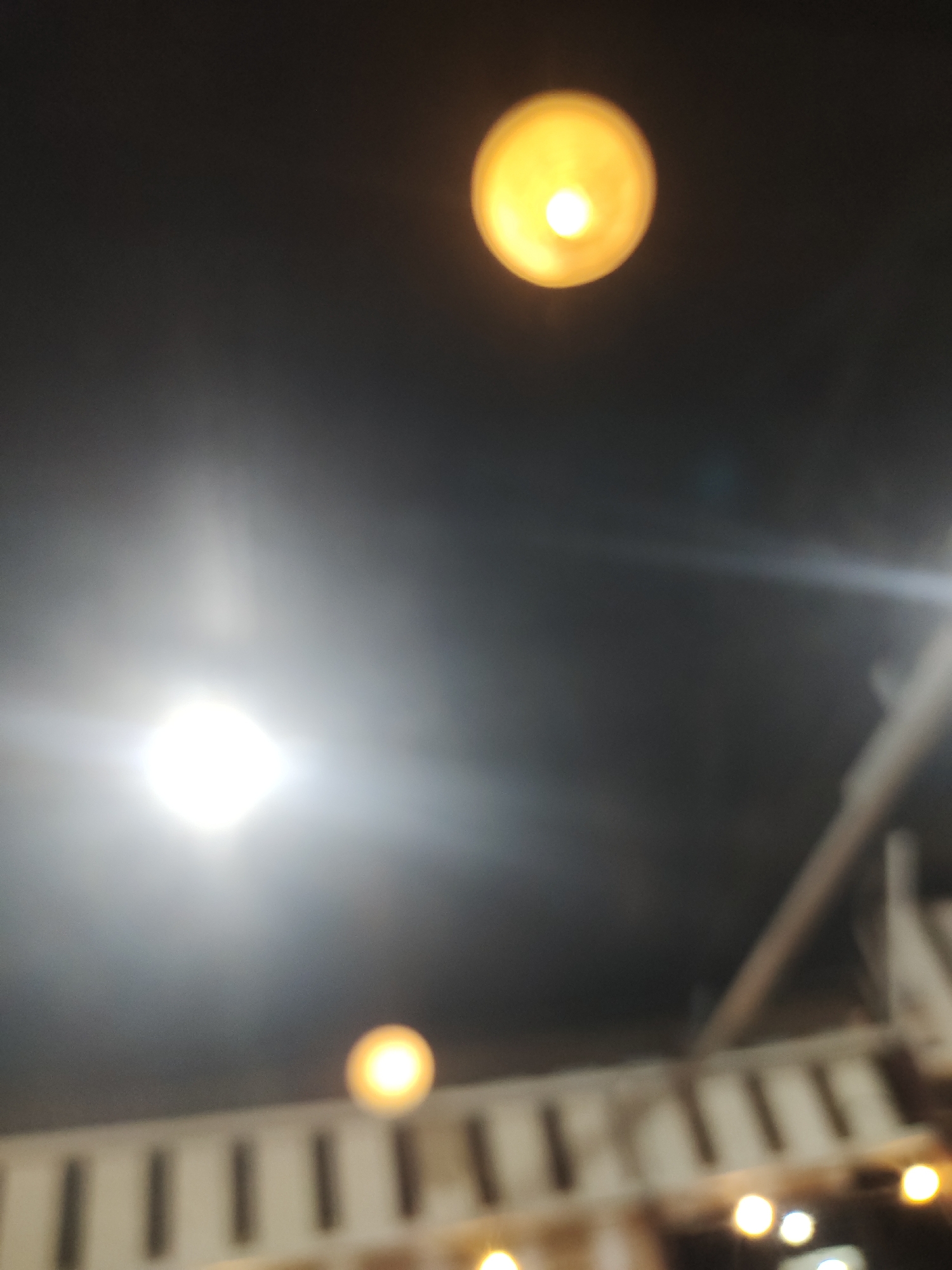 An out-of-focus, dimly lit indoor scene, captured in Keudematangkuli, Indonesia, features multiple artificial light sources against a dark background. Dominating the lower-left-to-central area is a very bright, white, somewhat square-shaped light, casting a significant glow. Two additional smaller, circular, yellow-orange lights are visible: one prominently in the upper-middle section, and another fainter one towards the bottom edge of the frame. Several other small, diffused light blurs are present at the very bottom.
The background, particularly on the right and bottom, shows blurry architectural elements, suggesting an interior structure like a ceiling or upper wall with repeating vertical features resembling beams or slats, and some diagonal supports. The entire image is blurred, creating a soft, bokeh effect around the lights, which are the main subjects. The dark environment indicates an interior setting, likely at night or in a space without natural light. No people or specific activities are depicted, and no text is visible.