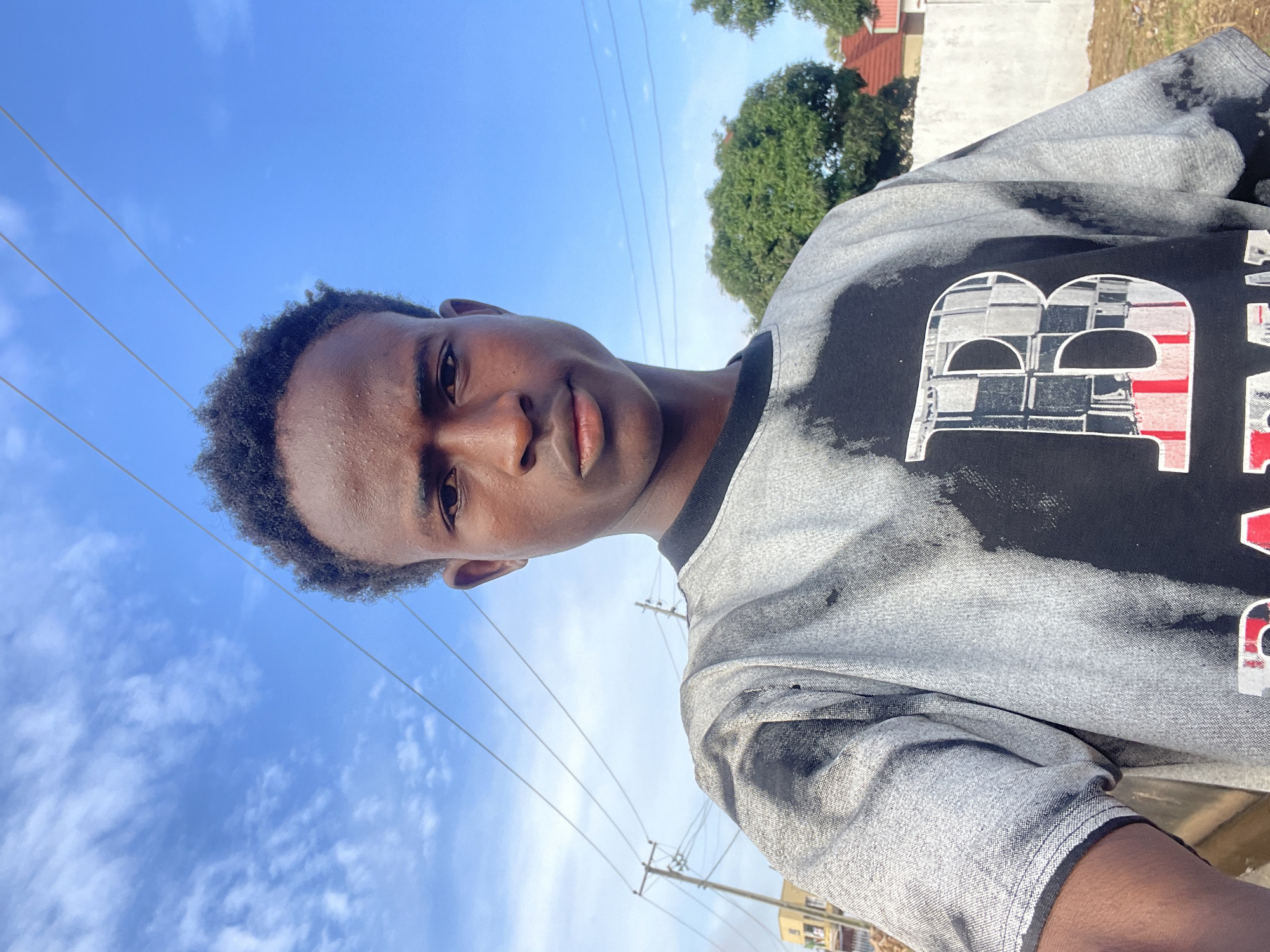 A young Black male is depicted in a close-up, chest-up selfie taken outdoors in what appears to be a residential area. The background features a clear blue sky with scattered clouds, overhead power lines supported by utility poles, and glimpses of buildings and trees. The subject is wearing a t-shirt with a distressed tie-dye pattern in black and gray, featuring a large stylized letter 'B' on the front. The subject's facial expression is neutral, with his gaze directed towards the camera. The lighting suggests it is daytime, with the sun illuminating the scene from the front and slightly to the side.