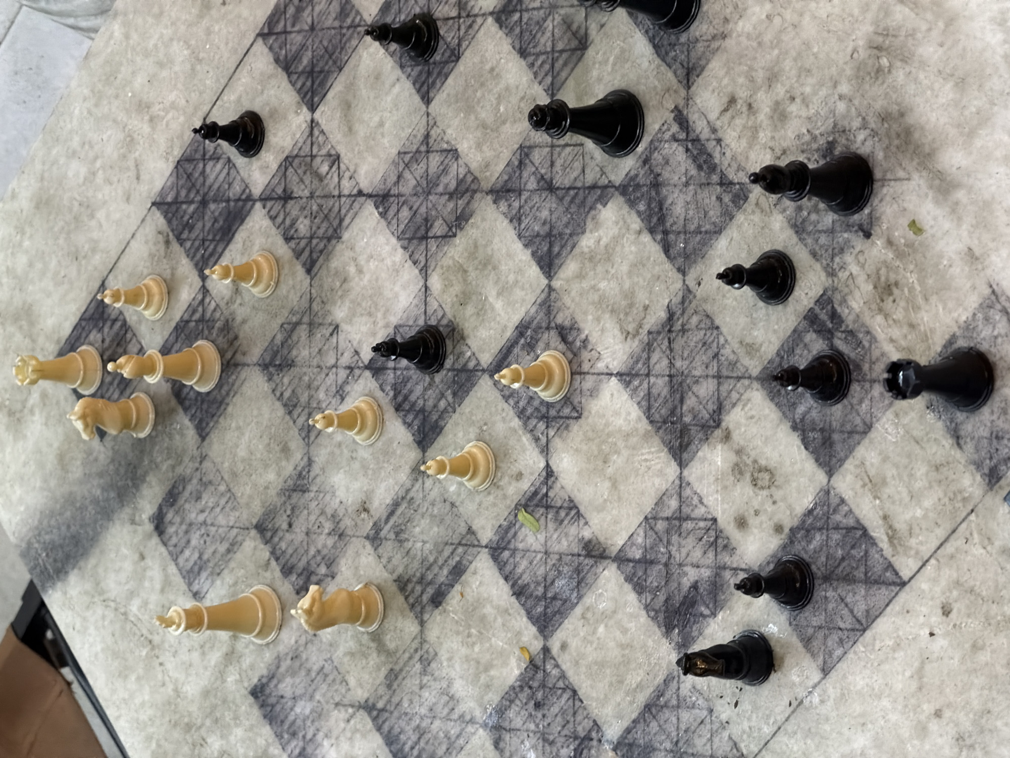 An overhead, slightly angled shot captures a unique chess game in progress on a distinctive board. The chessboard appears to be crafted from a light-colored, possibly white or light grey, polished stone or marble. The dark squares are not solid black but feature a textured, etched, or hand-drawn cross-hatch pattern in a darker grey or black, creating a rustic, artisanal look. The squares themselves are arranged diagonally relative to the frame.
Numerous chess pieces are scattered across the board, indicating a mid-game scenario rather than a starting position. On the left side, the white pieces, which have a yellowish, possibly aged or ivory-like appearance, include several pawns, a knight, a bishop, a rook, a queen, and a king. On the right side, the solid black pieces consist of several pawns, a knight, a bishop, a rook, a queen, and a king. The pieces are traditional Staunton-style designs, simple yet elegant.
The lighting is bright and even, suggesting natural daylight. The board shows some minor signs of use or specks of debris, adding to its authentic feel. There are no people visible, nor any specific location cues within the image itself. No text is visible anywhere in the frame.