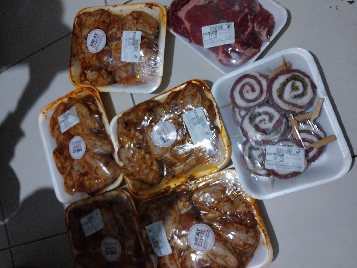 The image shows a collection of packaged meat products on a light-colored surface. There are six white styrofoam trays visible. Five contain what appears to be seasoned chicken wings, labeled with "MEAT ONE" stickers. The other two trays contain raw beef; one a cut of steak, and the other, meat rolls on skewers. All items are sealed in plastic wrap with price tags. The scene is possibly indoors, with a glimpse of tiled flooring and a shadow visible.