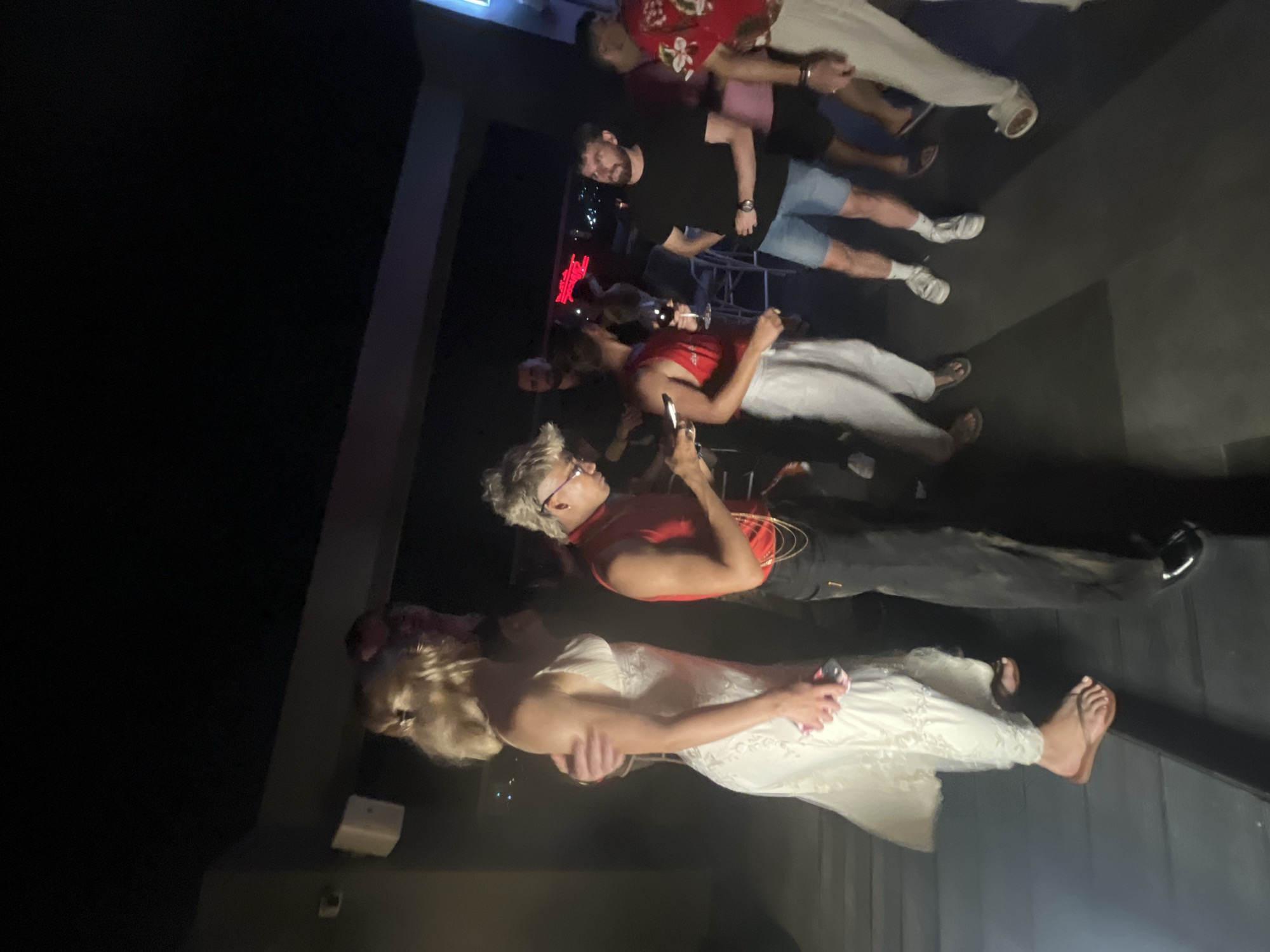 The media file depicts an indoor gathering of people. The scene is dimly lit, suggesting an evening or nighttime event, possibly a party or social function. Several individuals are visible, standing and interacting.
In the foreground, a person wearing a white, lacy dress is shown from the waist down, with their bare feet visible. They are holding a pink object in their left hand. To their left, another individual with blonde, short hair and glasses is wearing a red tank top and dark pants. They appear to be holding a phone.
Further in the background, a group of men are standing. One man wears a black t-shirt and denim shorts, holding a phone in his right hand. Another man is wearing a red shirt with a floral pattern. The overall atmosphere appears casual and relaxed.
The presence of drinks suggests a social event. The backdrop features dark, textured surfaces and some illuminated elements, including what appears to be a red neon sign in the distance. The location context provided is Đà Nẵng, Vietnam, but there are no specific visual cues within the image that definitively confirm this location. No legible text is visible in the image.