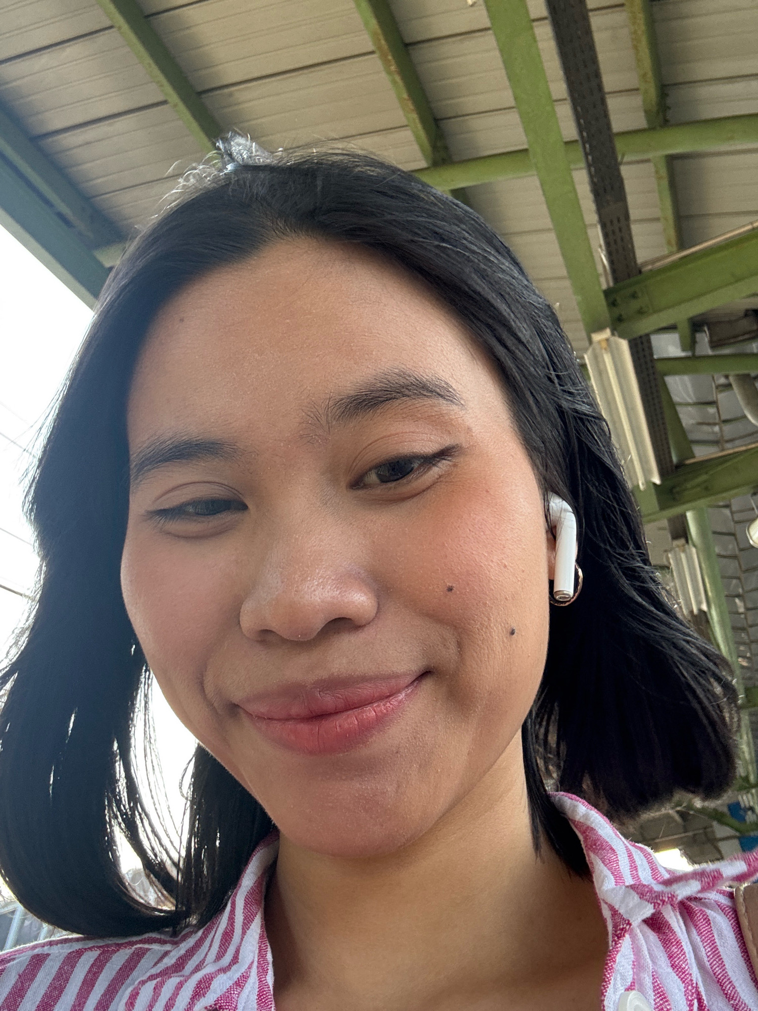 A close-up, low-angle selfie shows a young Asian woman with dark hair and light brown eyes. She is wearing a pink and white striped shirt and white earbuds. She has a faint mole on her left cheek and a subtle smile. The background is a slightly out-of-focus view of a building's interior, with green beams and corrugated metal roofing. The lighting suggests an outdoor or well-lit indoor environment. This scene could be anywhere in the city of East Jakarta, Indonesia, as there are no specific visual cues to pinpoint an exact location.
