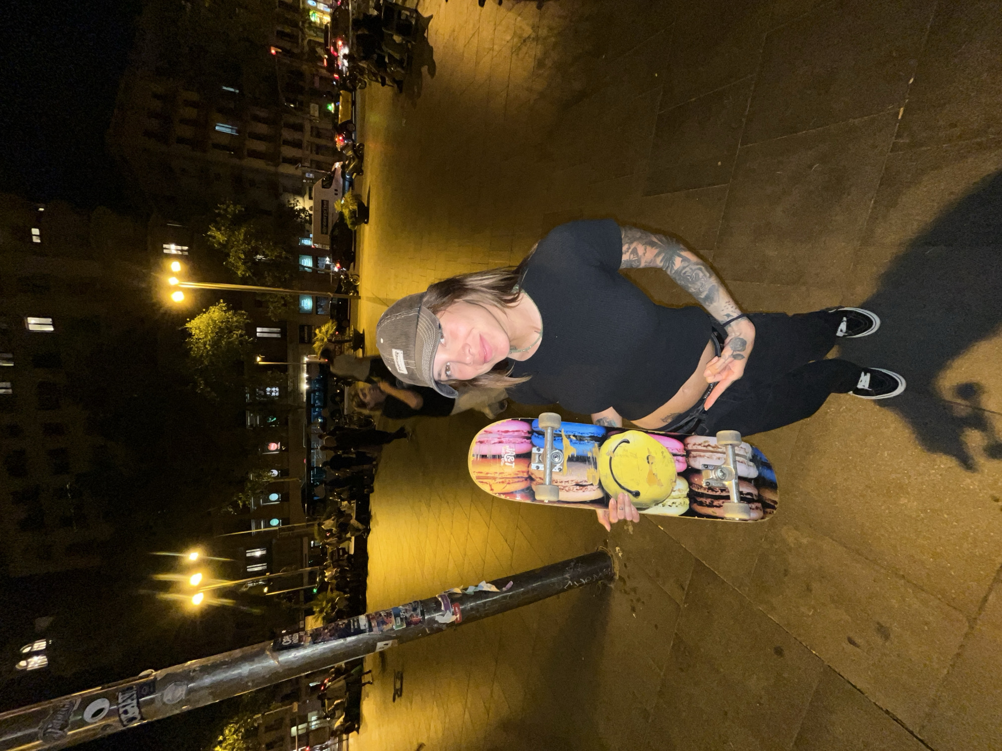 In an urban nighttime setting in Barcelona, Spain, a young woman with a skateboard stands on a paved plaza. She is wearing a dark t-shirt, black pants, and a grey baseball cap. Her arms are adorned with tattoos. The skateboard, held vertically in her right hand, features a colorful design of macarons and a prominent yellow smiley face sticker. She is looking towards the camera with a slight smile. The background reveals a street scene with people gathered and sitting on the ground, illuminated by streetlights and the lights of buildings. Vehicles, including cars and a van, are visible on the street. The lighting conditions suggest a late hour, with strong contrasts between illuminated areas and shadows. The overall atmosphere appears to be that of a public gathering or nightlife.