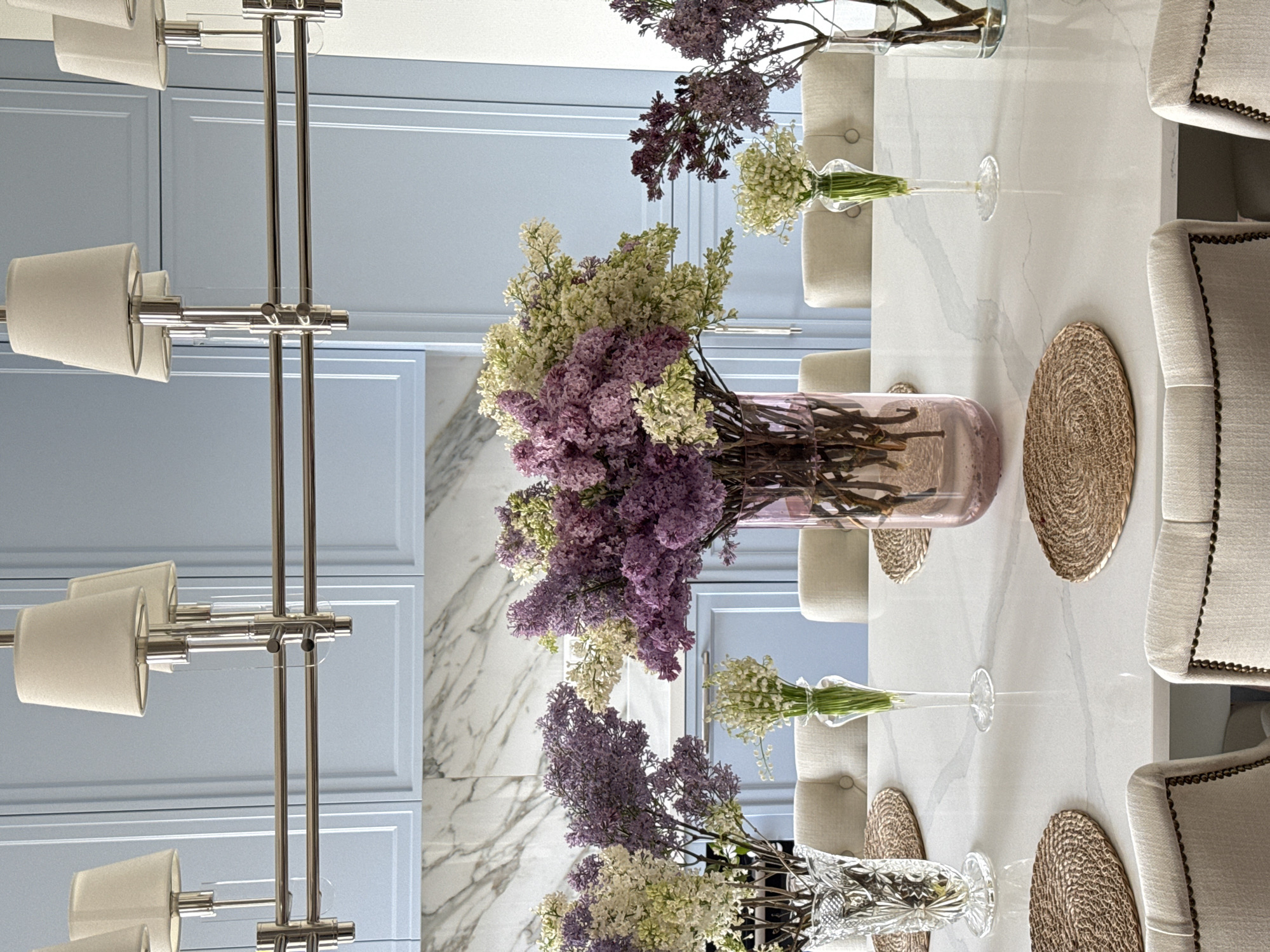 A dining table setting under a modern chandelier. Lilac flowers in various vases are arranged down the center of a white marble table. Place mats are set at each seat, which consists of light-colored chairs with dark trim. The setting is in an interior with light blue paneled walls. Location: Moscow, Russia is not determinable.