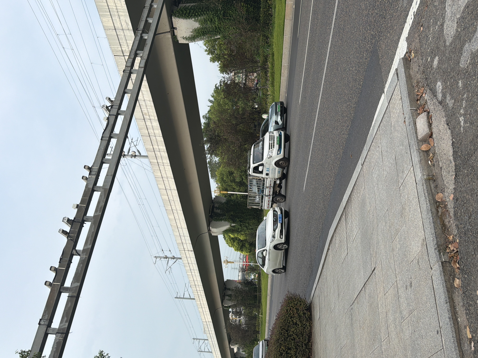 A road and sidewalk scene in Jiangjin, China, is depicted under an elevated structure, likely part of a transportation system such as a railway or highway. The elevated structure, supported by concrete pillars covered in ivy, spans across the image horizontally, with visible overhead wires and cameras mounted on its underside. On the road, a white sedan, a white truck with a flatbed trailer, and a light green sedan are visible. The white sedan is in the left lane, followed by the truck, and then the green sedan in the right lane. The sidewalk, made of gray paving stones, is to the left of the road, bordered by a low hedge. The sky is overcast. The scene appears to be a typical urban or suburban environment with traffic and infrastructure.