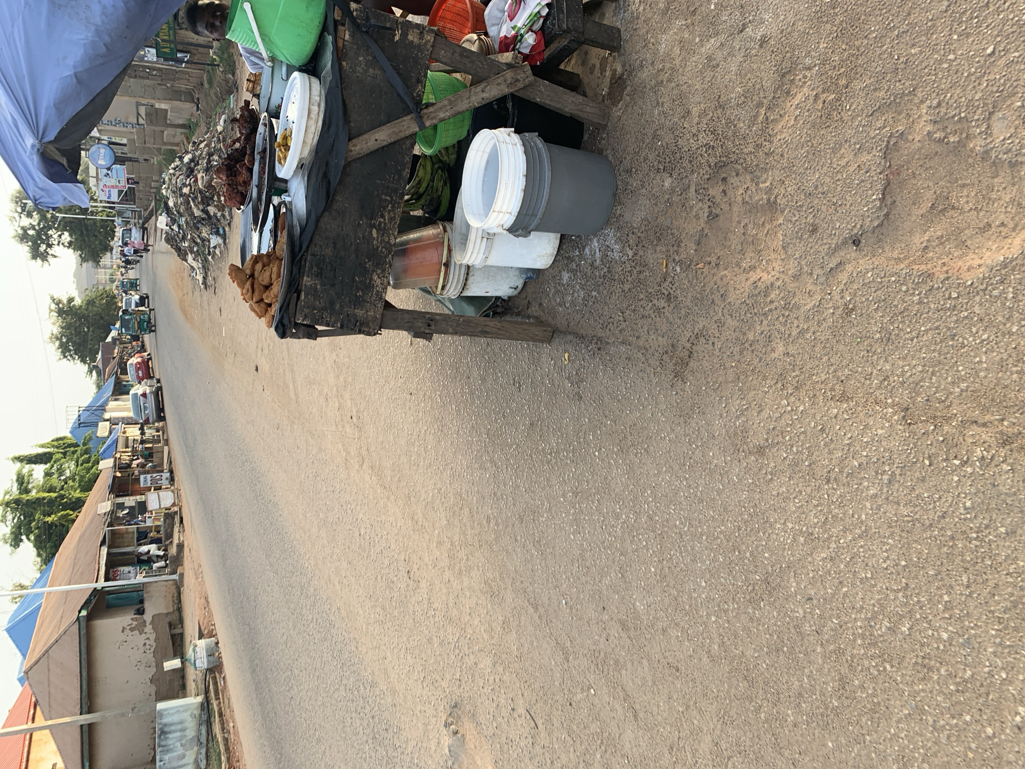 A street scene in New Stadium, Nigeria, shows a food vendor's stall positioned on the side of an asphalt road. The stall, constructed from wood, has a covering made of black fabric and holds several buckets and containers, along with trays of prepared food items. These include fried foods in varying shapes and colors. The stall is under a blue canopy, and a person is partly visible behind the stall.
The asphalt road shows signs of wear. A line of buildings, mostly one-story structures with metal roofs, runs along the opposite side of the road. A variety of shops and businesses are visible, and there are people present along the street. Vehicles, including cars and commercial transportation, are moving along the road in the distance. Some vegetation, including trees, is visible in the background.