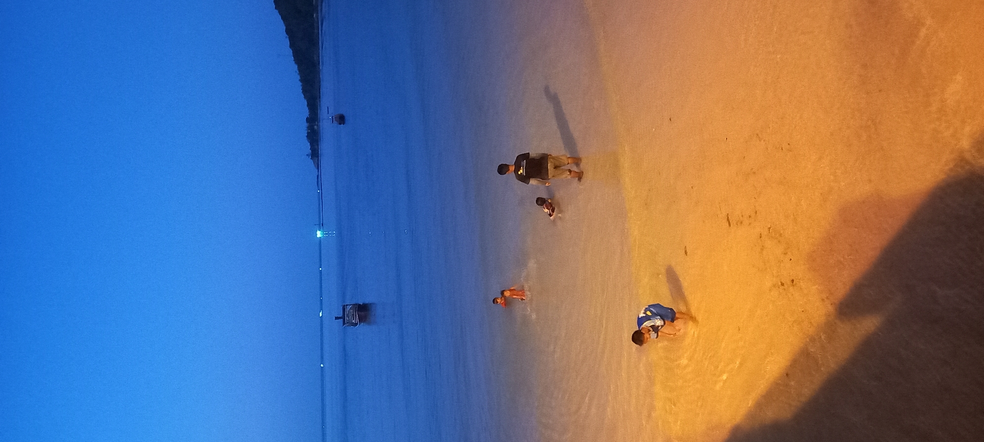 An evening scene unfolds on a beach, likely at dusk or night, illuminated by prominent artificial light. The sky, visible in the upper left, is a deep, uniform blue, indicative of low light conditions. The image is split diagonally by contrasting light; the upper left half consists of the dark blue water and sky, while the lower right half shows shallow water and sand brightly lit with a warm, orange glow.
Four individuals are present in the shallow water near the shoreline. An adult, wearing a dark top and light shorts, stands closer to the transition between the illuminated and darker water. Two children are nearby; one wearing a dark top is close to the adult, and another in an orange-colored swimsuit or top is further into the water. Closer to the foreground right, a fourth child, dressed in a blue and white top, is crouching or playing in the brightly lit shallow water. All figures cast long shadows due to the strong directional lighting, which emanates from the right side of the frame.
In the mid-ground, extending into the darker blue water, several boats are visible, some with small lights. A long, narrow structure, possibly a pier or jetty, also stretches across the water with intermittent lights, leading towards what appears to be a forested coastline in the far distance. The mood is calm and relaxed, suggesting recreational activity at the beach during the evening. No visible text is present in the image. The elements, particularly the boats, align with the provided location context of Baan Chaloklum, Thailand.