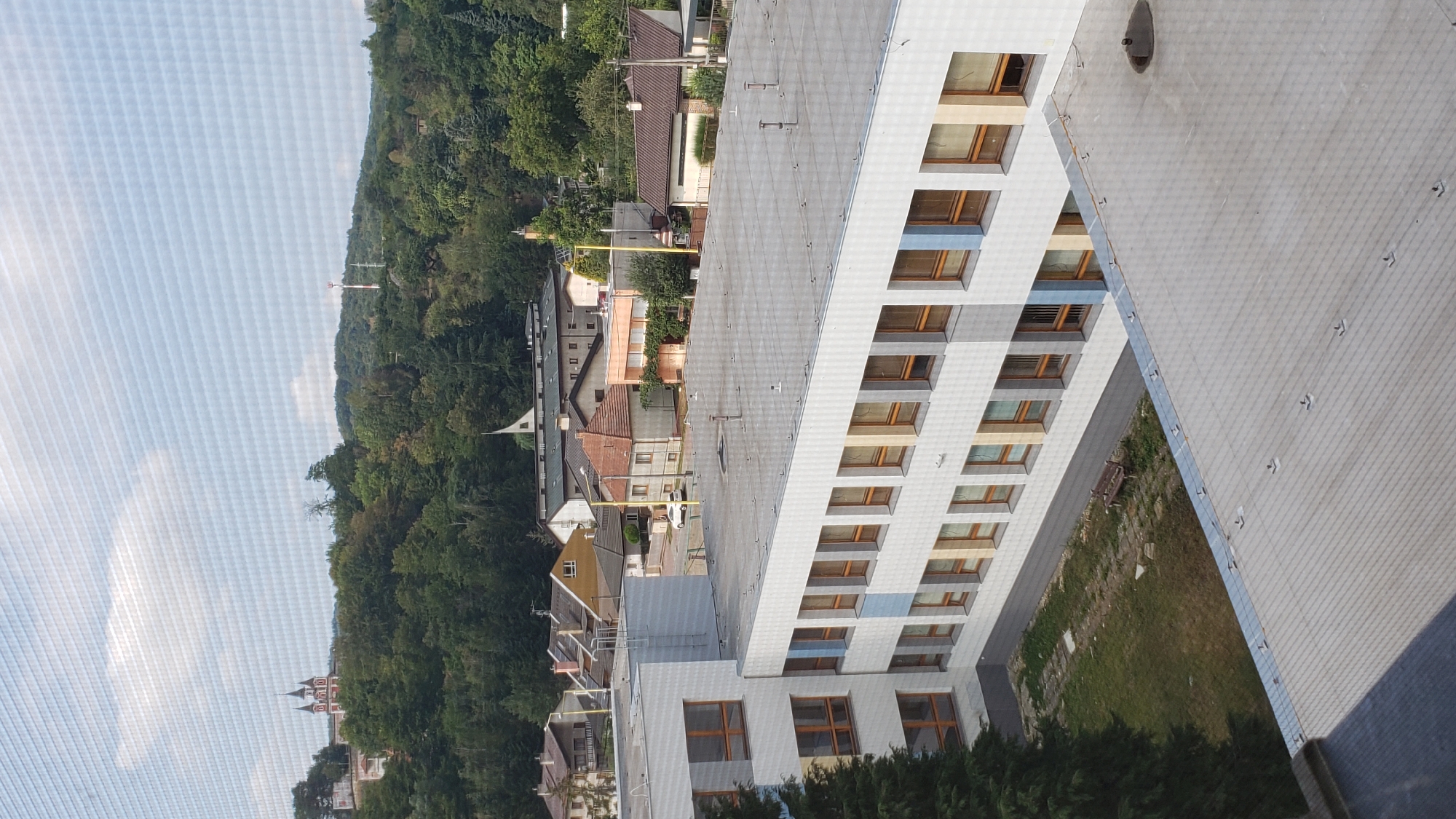 The image shows a view from a high-rise building, looking out over a suburban area and surrounding hills. In the foreground, a modern, multi-story building with a light-colored facade and numerous windows is visible. A small courtyard with a grassy patch, a paved path, and a wooden bench is located next to this building. Beyond the foreground building, a collection of smaller residential houses with pitched roofs are scattered across the landscape. In the distance, a lush green forest covers the rolling hills, and a prominent church with red and white architecture and spires is situated on a hilltop. A cell tower is also visible on another hill in the background. The sky is partly cloudy, with a hazy appearance, possibly due to a screen or filter. The overall scene depicts a residential area with natural surroundings in Prešov, Slovakia.
