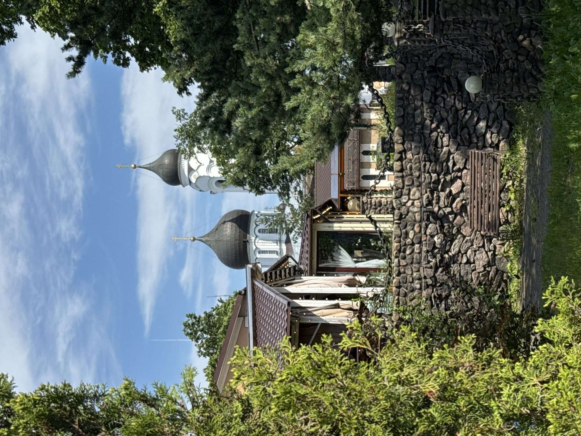 The image displays an outdoor scene in Lomonosov, Russia, with a focus on architectural elements and natural greenery. In the background, the domes of a white church with dark grey onion domes pierce the bright blue sky, which is dotted with wispy white clouds and a faint contrail from an airplane. The church's crosses are visible at the apex of the domes.
In the foreground and mid-ground, the scene is framed by lush green trees and bushes, some of which appear to be cypress or juniper. Partially obscured by this foliage is a building with a reddish-brown, textured roof. Adjacent to this building is a distinctive structure built with a rough, dark grey stone wall, possibly a decorative fence or part of a larger construction. This stone wall is adorned with a heavy, dark metal chain that wraps around it and hangs down. A simple, rustic metal bench is positioned near the base of the stone wall. There are also visible light fixtures, possibly string lights, illuminating some of the structures.
The overall impression is one of a sunny day, with clear skies suggesting daytime. The style of the church architecture, particularly the onion domes, is characteristic of Russian Orthodox churches. The combination of historical architecture and natural elements creates a serene and picturesque setting. No people are visible in the frame, and there is no visible text. The composition suggests a deliberate framing of the church and its surroundings, with the natural elements leading the viewer's eye.