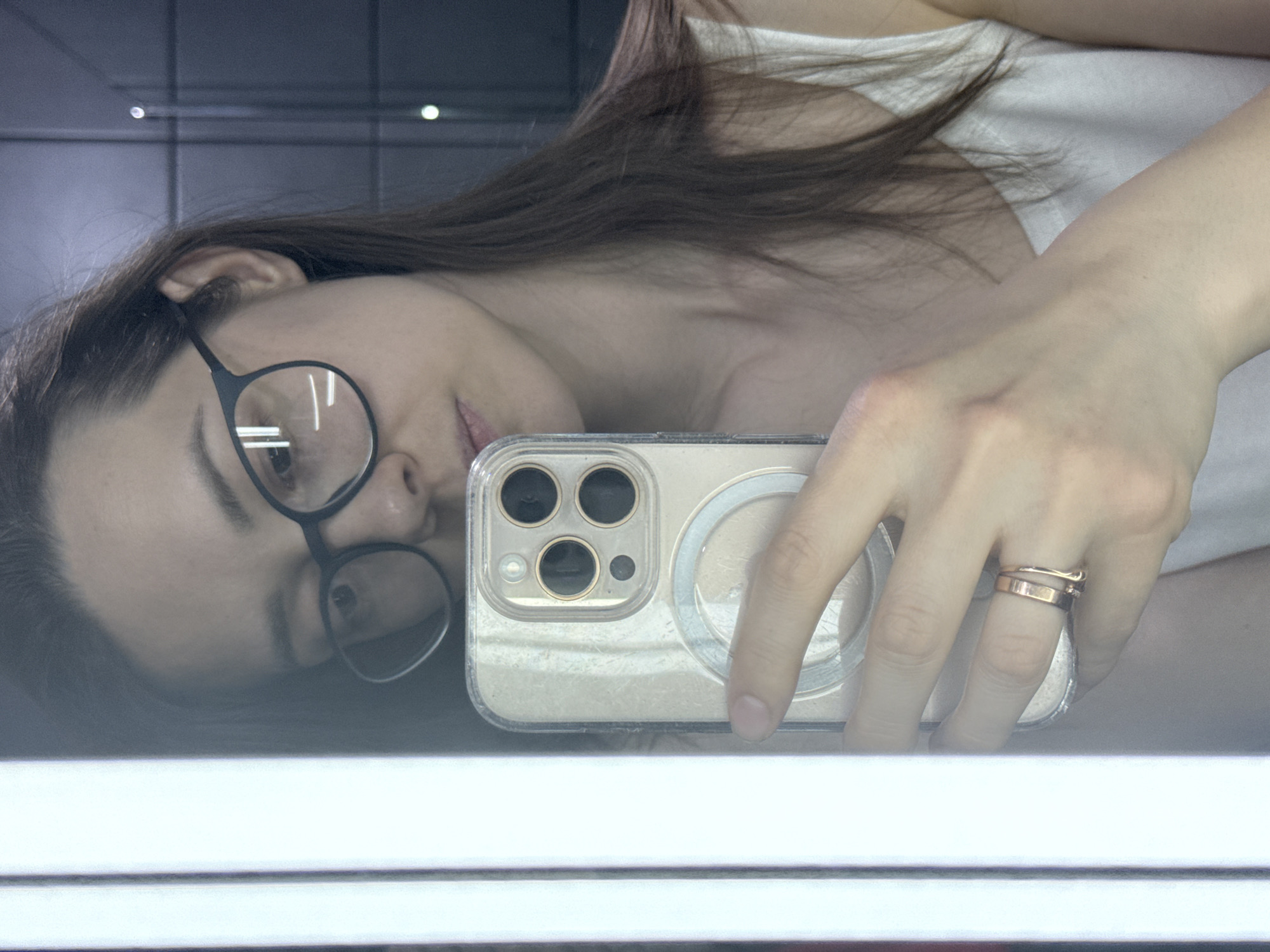 The image displays a mirror selfie of a female individual from the chest up. She wears a white tank top and black-rimmed glasses. Her long, dark brown hair frames her face. Her right hand, adorned with two gold-toned rings on the ring finger (one a simple band, the other a thinner, possibly wavy design), holds a gold-colored iPhone in a clear MagSafe-compatible case. The phone's triple camera system is clearly visible. The woman's gaze is directed towards the smartphone's camera lens. A prominent vertical strip of bright white light is reflected on the left side of the mirror. The background, visible behind the woman in the reflection, consists of a dark, possibly tiled or paneled wall with a faint grid pattern and subtle circular light reflections in the upper right quadrant. The specific location within the city of Chaikovsky, Russia, cannot be determined from the visual evidence presented in the image.
