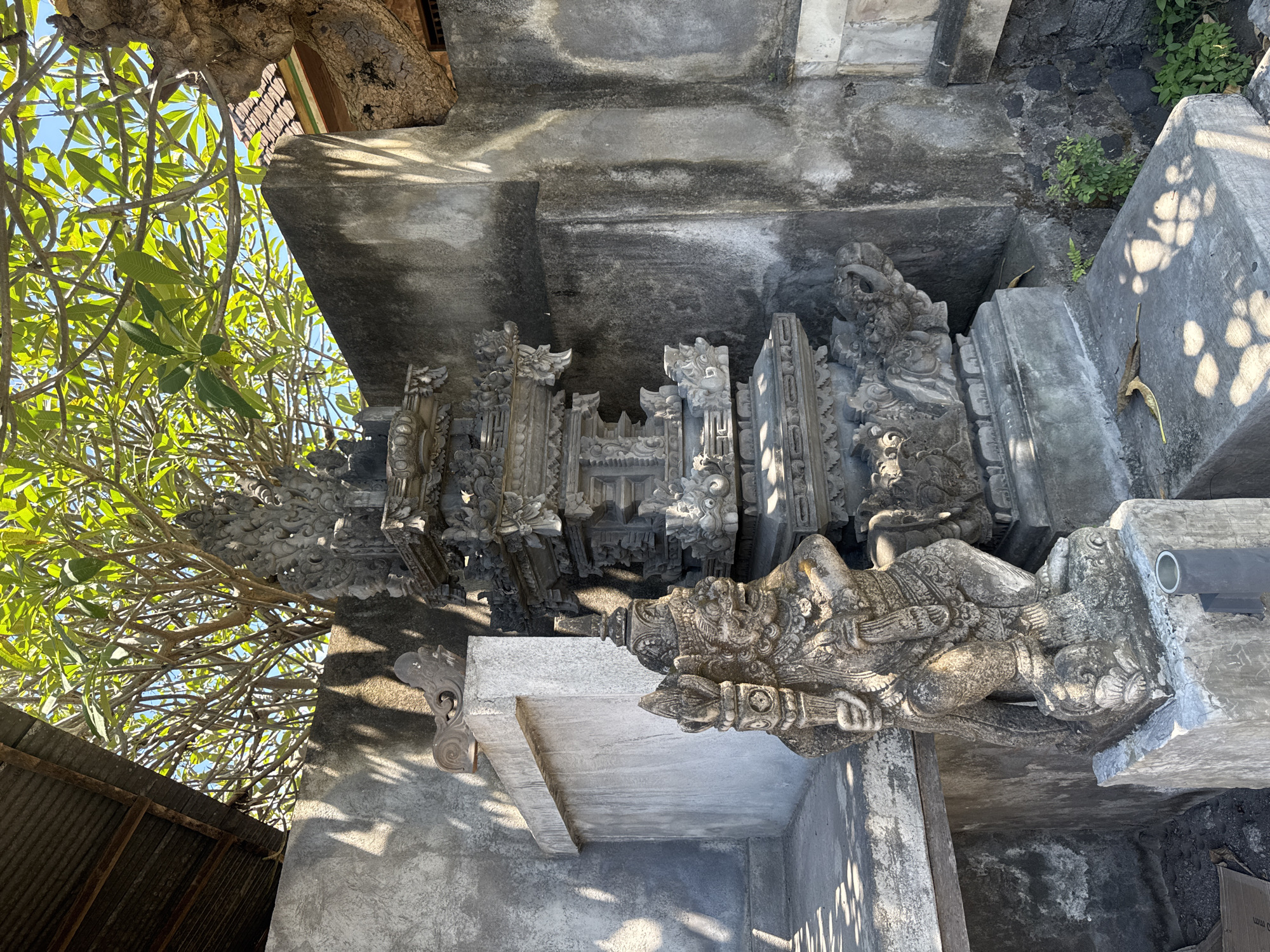 The image depicts a stone structure in Kubu, Indonesia, possibly part of a temple or religious site. The architecture features intricate carvings and sculptures, suggesting a traditional Balinese style. Lush green foliage and trees are visible in the background, indicating a tropical environment. The structure appears weathered and aged, implying its historical significance. There are no people visible in the scene. The image suggests a cultural or religious landmark, likely maintained as a historical or spiritual site.