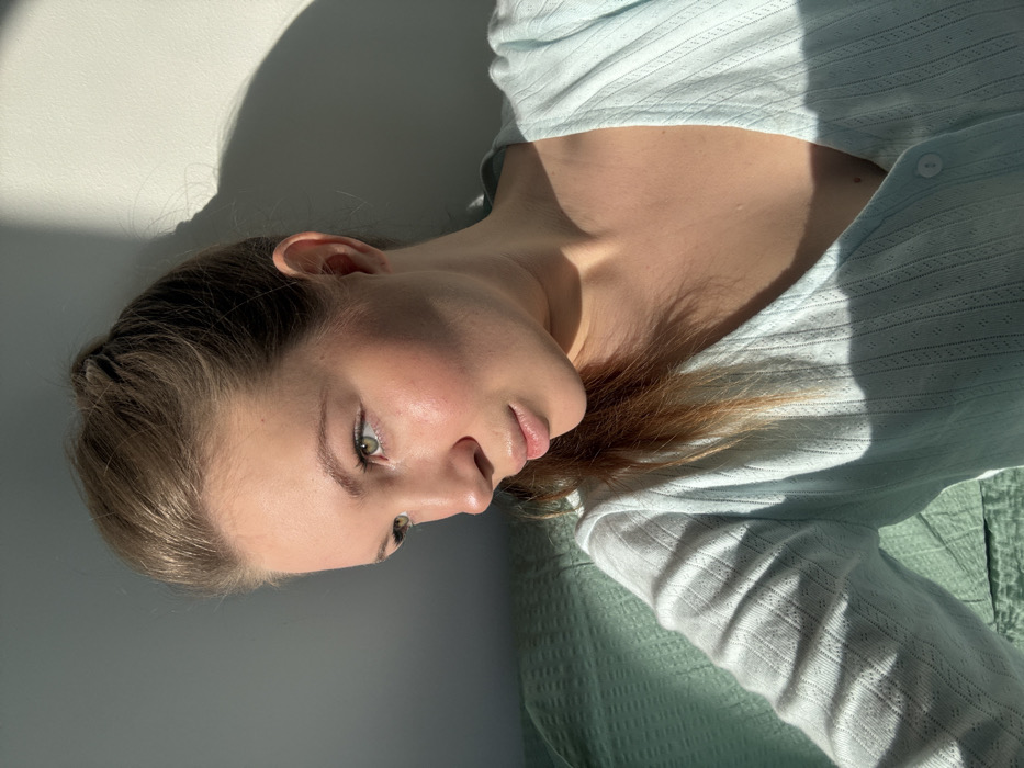 An indoor photograph features a female subject, depicted from the chest up, positioned within a residence in the city of Vozdovac Urban Municipality, Serbia. The subject has fair skin, green eyes, and light brown hair tied back. She wears a light blue, V-neck, ribbed cardigan with a perforated pattern. Strong, directional sunlight illuminates the left side of her face and body, casting distinct shadows on the light-colored wall behind her and across her right side. A green, textured fabric object is visible in the lower left quadrant of the frame and behind the subject's left shoulder. The subject is gazing towards the left of the frame. No other individuals or specific actions are observed.