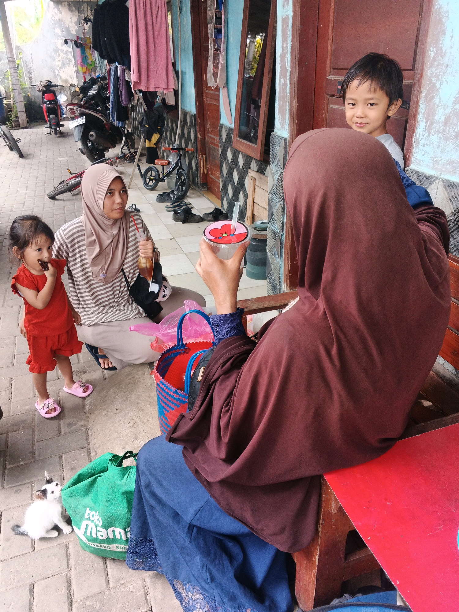 In Kadu, Indonesia, a woman wearing a brown hijab and a striped shirt crouches on a paved walkway, holding a plastic cup with a red straw. Beside her, a young girl in a red outfit stands, holding a piece of chocolate to her mouth. A young boy in a grey shirt stands near a wooden door, looking towards the viewer. A small white cat with grey markings sits near a green tote bag with "toko mama" printed on it. In the background, motorcycles, bicycles, and laundry hang from a line. The scene appears to be an outdoor area in front of a building with a light blue wall and wooden doors.