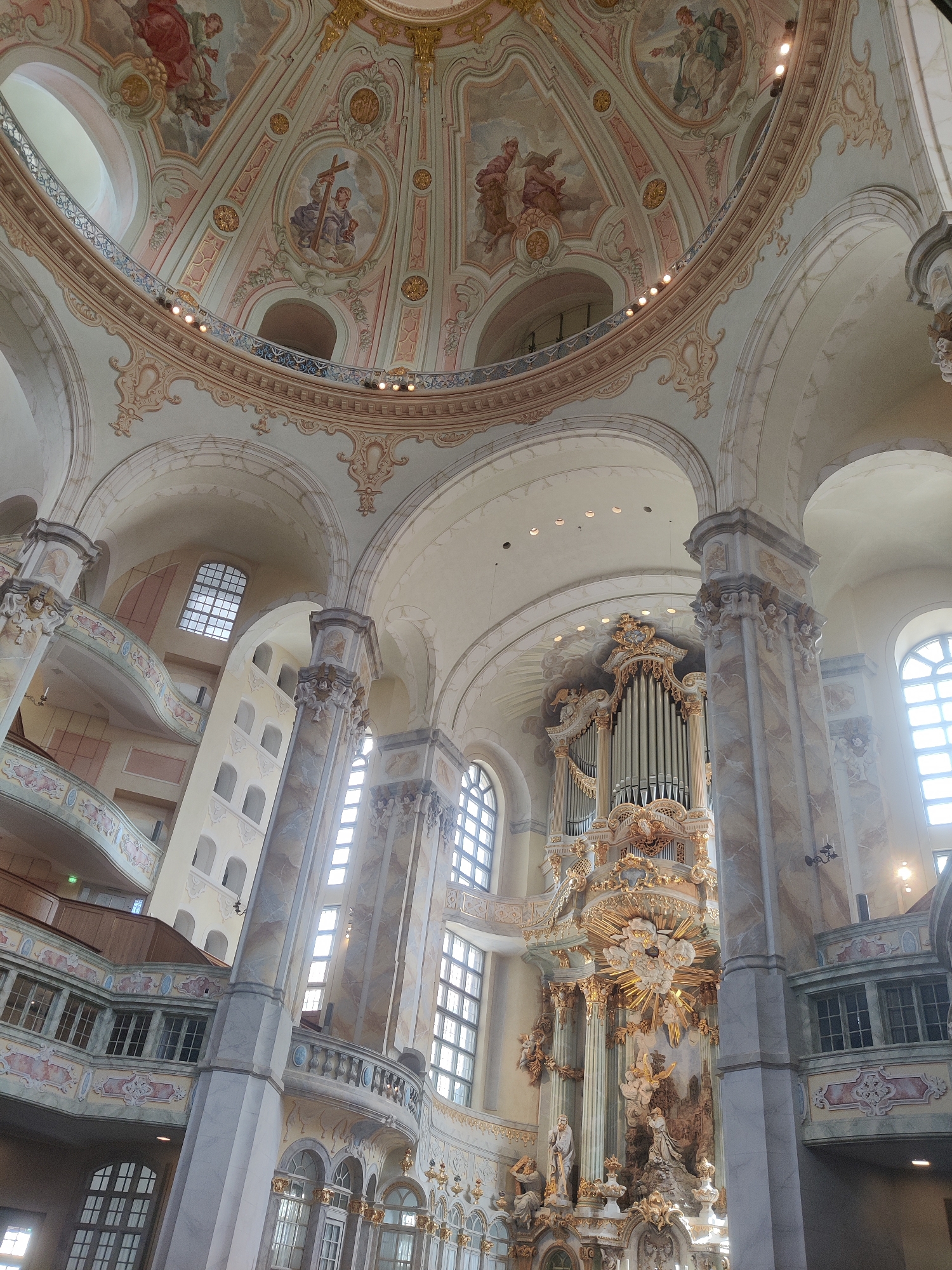 The image shows the interior of the Frauenkirche church in Dresden, Germany. The perspective is from the ground floor looking up towards the dome and a grand organ. The dome is elaborately decorated with frescoes depicting religious scenes and ornate architectural details in pastel colors. A circular balcony with decorative railings runs beneath the dome. To the right of the dome, a large, gilded pipe organ with intricate carvings and sculptures is prominently displayed. The walls of the church are adorned with marble-like columns and painted details, and large arched windows allow natural light to enter. Several levels of balconies with railings and windows are visible on the side walls, indicating the multi-tiered structure of the church's interior. The overall impression is one of grandeur and ornate Baroque architecture.