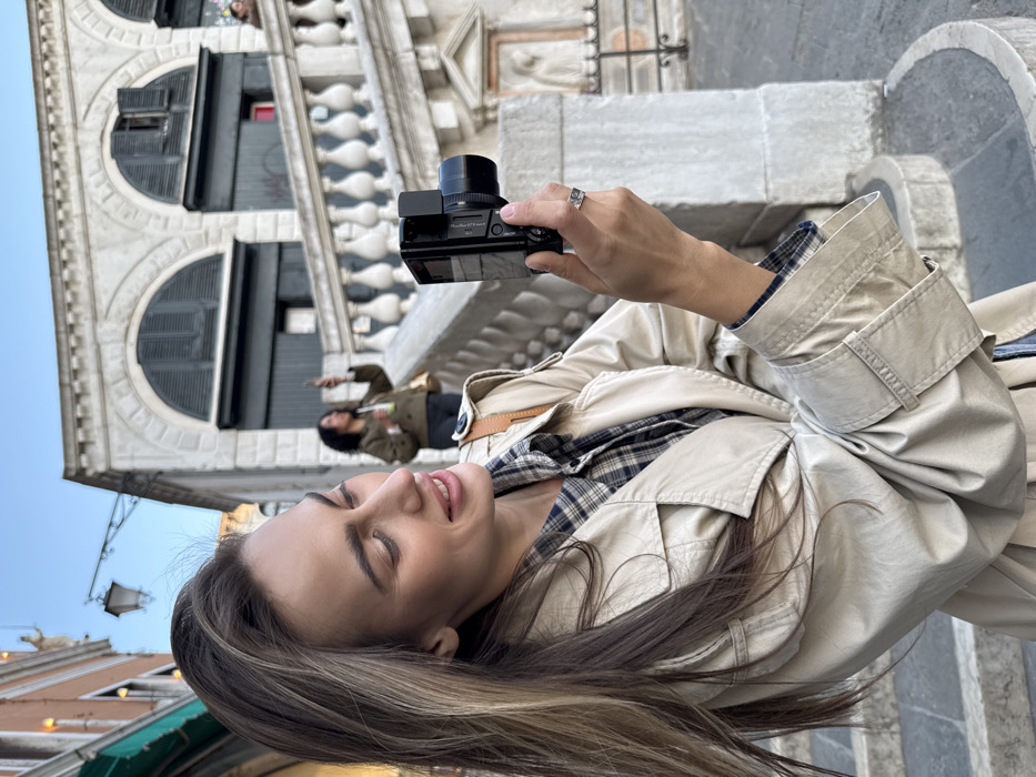An outdoor, eye-level shot depicts a woman in the foreground smiling while holding a black compact digital camera in her right hand, positioned to her right. She has long brown hair with blonde highlights, styled down, and wears a light beige trench coat over a blue and white plaid shirt. A silver-colored ring adorns her right ring finger. A light brown strap from a bag is visible over her right shoulder.
The scene is set in Venice, Italy, near the Rialto Bridge (Ponte di Rialto). In the midground, the distinctive light-colored stone balustrade and staircase of the Rialto Bridge ascend from left to right. Archways with dark shutters are visible beneath the bridge structure. Further up the staircase, a second woman with dark, curly hair, wearing a dark olive-green jacket, is observed holding a smartphone aloft with both hands, seemingly taking a photograph. She also holds a light-colored cup or bottle in her left hand.
To the far left of the frame, a building with an orange-brown facade and a small statue on a ledge is visible. An ornate black lamppost extends from this building towards the center. The background features a clear, bright blue sky. Stone steps and architectural details of the bridge are prominent throughout the scene.