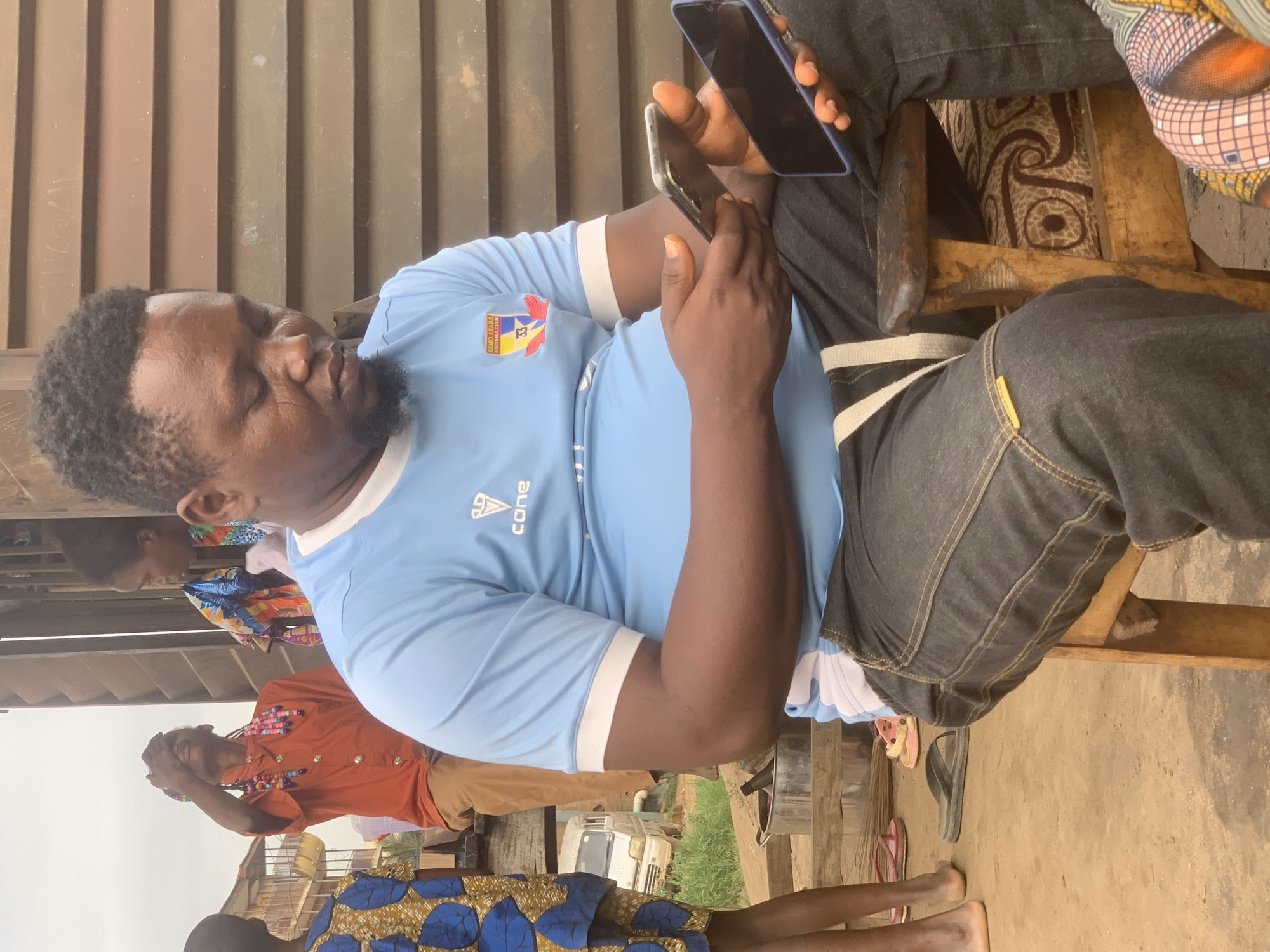 In Shagamu, Nigeria, a man wearing a light blue t-shirt with the "Remo Stars Football Club" logo and dark jeans sits on a wooden stool. He is holding two smartphones, one in each hand, and appears to be looking at the phone in his right hand. His gaze is directed downwards. Behind him, a wooden wall with horizontal planks forms the background. To his left, a young child wearing a blue and yellow patterned garment stands, and further left, another person in an orange shirt and brown pants is visible. Several pairs of flip-flops are scattered on the dusty ground in front of them. A white van is parked in the background, and various household items are present. The scene suggests a casual, outdoor gathering.