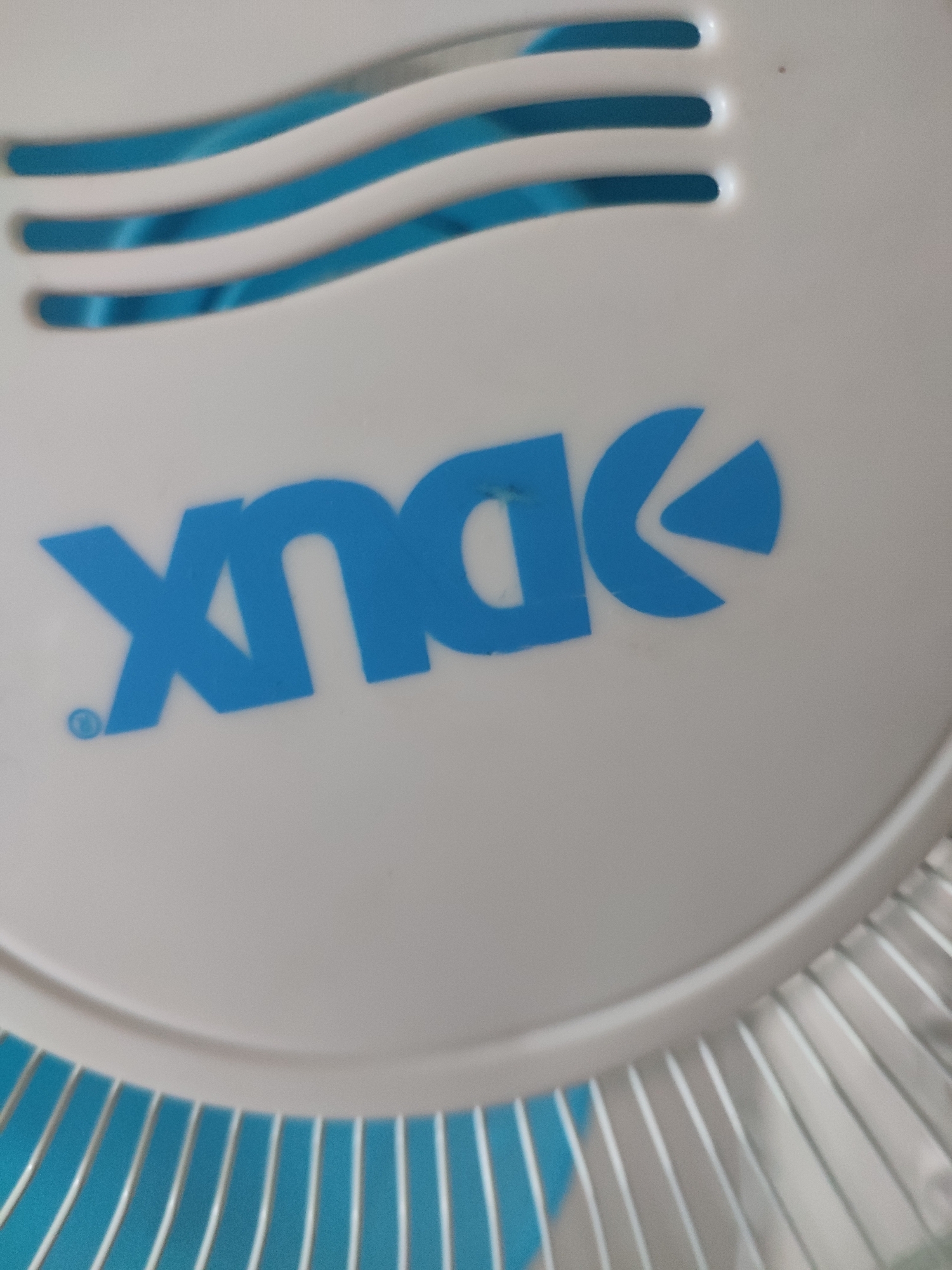 The image is a close-up shot of a white fan with a blue logo. The logo appears to be stylized text, with the letters "X N O" or similar visible, followed by a symbol resembling a curved arrow or a crescent moon shape. The letters are in a bright blue color, and the "X" has a small blue circle next to it, possibly representing a registered trademark. Above the logo, there are three horizontal blue stripes on the white surface, suggesting a design element or part of the fan's grill. The fan is likely indoors, given the lighting and the absence of any outdoor environmental cues. The bottom of the image shows a portion of the fan's grill, with thin metal bars against a bright blue background. The overall impression is of a product shot focusing on the branding of the fan. No specific time of day, weather conditions, or emotional cues are discernible. The location context provided is Naroŭlia, Belarus, but there are no visual elements within the image that directly confirm this location.