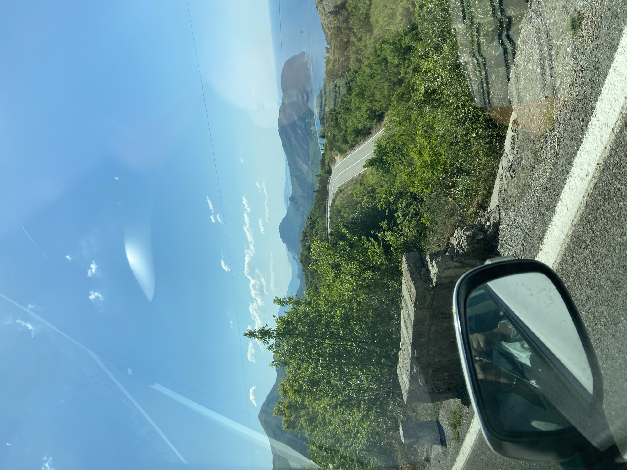 The image captures a view from inside a vehicle, likely a car, traveling on a mountain road. The location is suggested by the query to be in the city of Nerin, Montenegro. The road winds through a mountainous landscape with lush green vegetation. In the distance, there's a view of the sea. The sky is clear and blue with a few scattered clouds. The car's side mirror is visible in the foreground, reflecting the interior of the car. The road has a white line marking the edge. The image has a slight upward angle and appears to have been taken through a window, as evidenced by the reflections and artifacts in the blue sky.