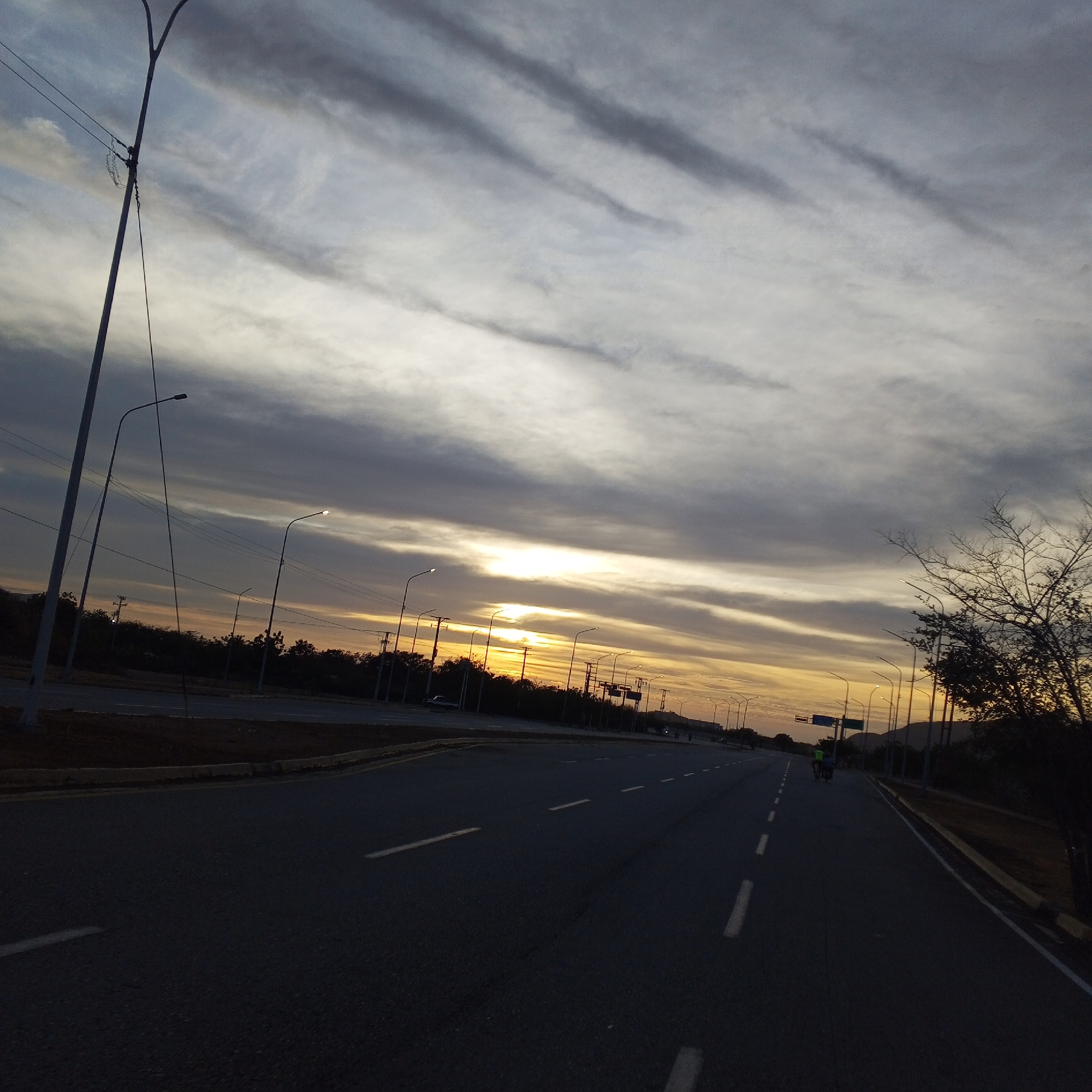 The image depicts a road at sunset in Altagracia, Venezuela. The road is divided by white dashed lines, with streetlights lining both sides. A person on a bicycle can be seen in the distance on the right side of the road. The sky is filled with clouds, with a warm sunset glow visible behind them. On the left side, there is a secondary road separated by a median strip with vegetation. Bare trees and utility poles are visible on the right edge of the frame.