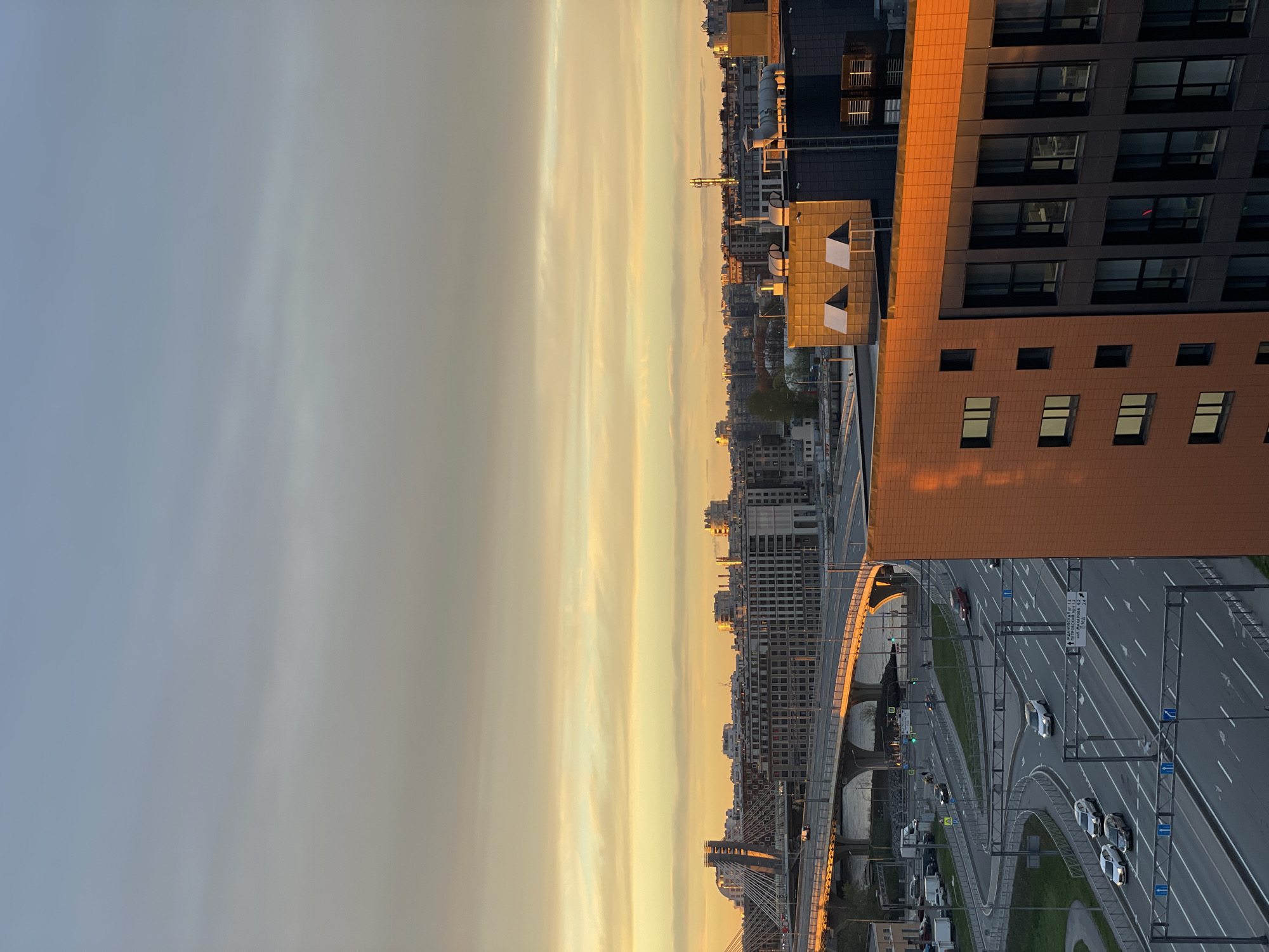 The image shows a high-angle view of a cityscape during sunset or sunrise. In the foreground is a multi-lane highway with several cars. To the right, a tall, modern building with windows is visible. In the distance, a bridge arches over a river or road. Numerous buildings fill the cityscape, and the sky is a gradient of colors from grey to orange. The location is Saint Petersburg, Russia, indicated by the street signs written in Russian.