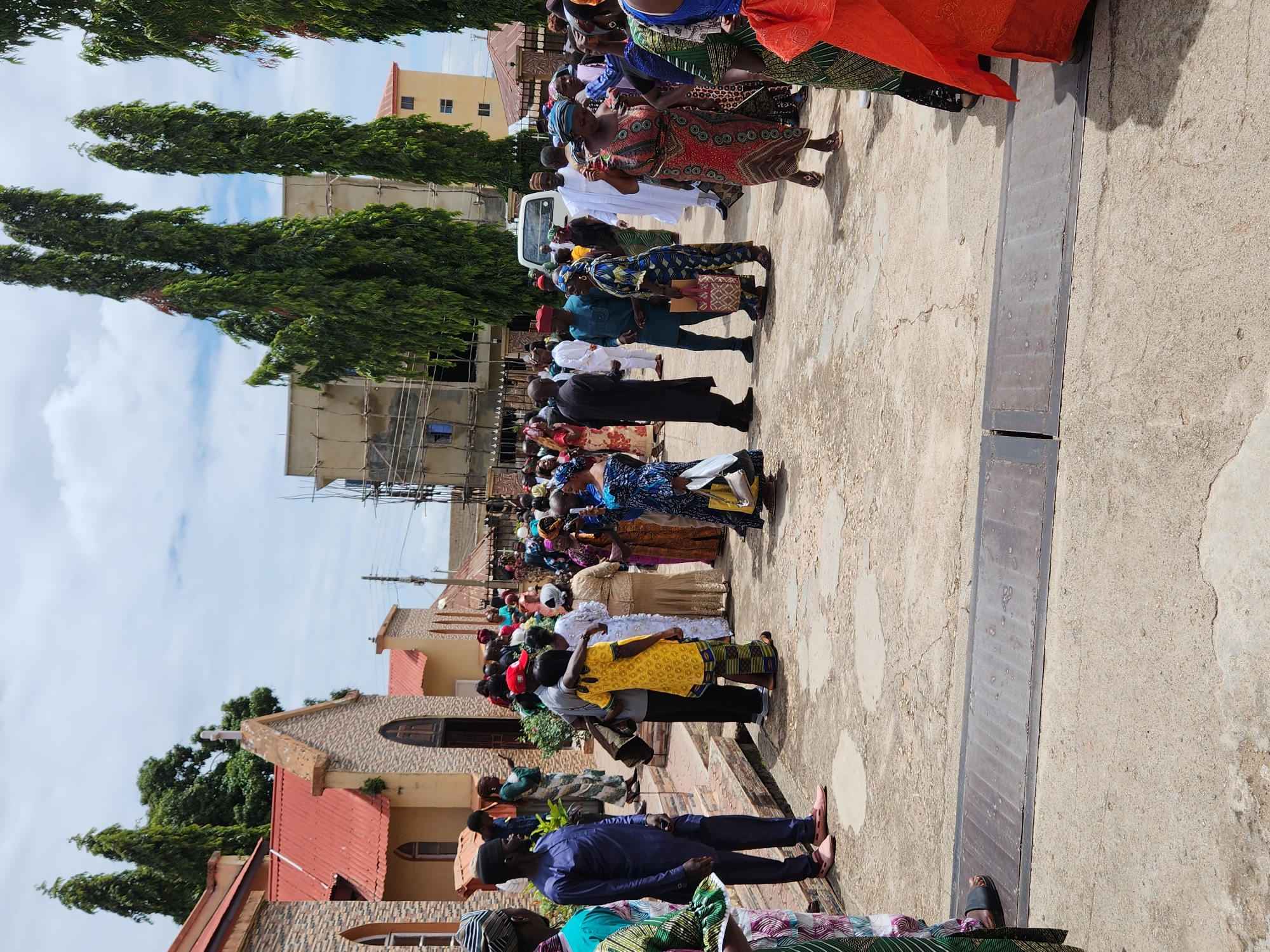 The image captures a bustling outdoor scene featuring a large gathering of individuals, predominantly adults, on a paved surface. The location is an open area in the city of Unknown, Unknown.
In the left background, a stone-clad building with a red corrugated metal roof and arched windows is visible, identified as a church by a white cross at its apex. Adjacent smaller structures with similar roofing are also present. To the right of the central gathering, a multi-story building under construction with visible scaffolding is partially obscured by tall, slender evergreen trees. Further right, another light-colored, multi-story building with multiple small windows is discernible.
The ground is a light-colored, cracked paved surface, possibly concrete, with a long, dark metal grate embedded horizontally across the foreground.
Numerous people, estimated to be over fifty, are distributed across the scene. They are dressed in diverse colorful attire, including patterned fabrics, traditional garments, and headwraps, suggesting a celebratory or formal social occasion. Many are engaged in conversation, standing in small groups, or walking.
Specific individuals include:
- A man in a dark blue outfit and light pink shoes standing towards the left foreground.
- A man in a red cap embracing a woman in a yellow patterned dress in the mid-ground, left of center.
- A woman in a red and blue patterned dress is walking towards the right foreground.
- A white van is partially visible behind the crowd on the right side of the mid-ground.
The sky is partially cloudy with patches of blue visible, indicating daytime conditions.