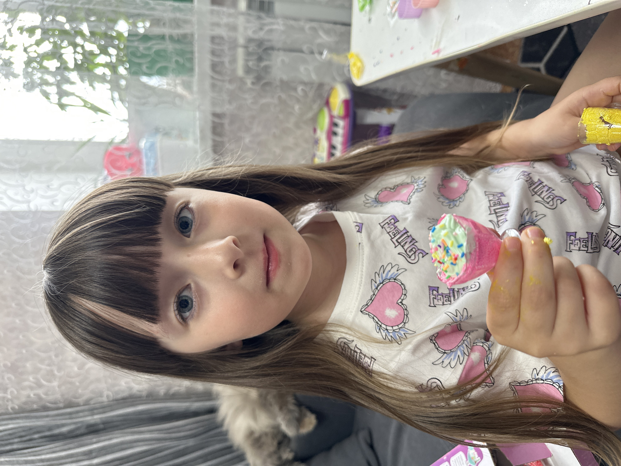 A young girl, approximately 5-8 years old, is depicted indoors, gazing directly forward. She has long, straight dark brown hair with a full fringe (bangs) across her forehead, accented by lighter streaks within the dark hair. Her eyes are a clear blue. She is wearing a white t-shirt featuring multiple pink winged heart designs and stylized purple text.
In her extended right hand, she holds a small, pink, stemmed goblet-like object. The goblet is topped with a light green, textured substance resembling whipped cream or soft-serve, generously covered with colorful sprinkles in shades of blue, yellow, orange, pink, and white. Traces of yellow residue are visible on her right thumb and index finger, suggesting recent handling of a colored material. A second, smaller, clear goblet containing a yellow, granular substance is partially visible in her left hand, positioned towards the bottom right of the frame.
The background to the girl's left features a dark grey upholstered sofa, on which a fluffy, light grey cat is resting, partially obscured and out of focus. Behind the sofa, grey patterned curtains are visible. To her right, a brightly lit window is covered by white, patterned sheer curtains, through which diffuse outdoor light and the faint outline of a green potted plant are discernable. Below the window, a white radiator is present. A white child-sized table, cluttered with small, colorful crafting items including pink containers and loose sprinkles, is positioned to her right. Further in the background, a vibrant purple, yellow, and green toy keyboard is visible, suggesting a play area. The walls appear light-colored.