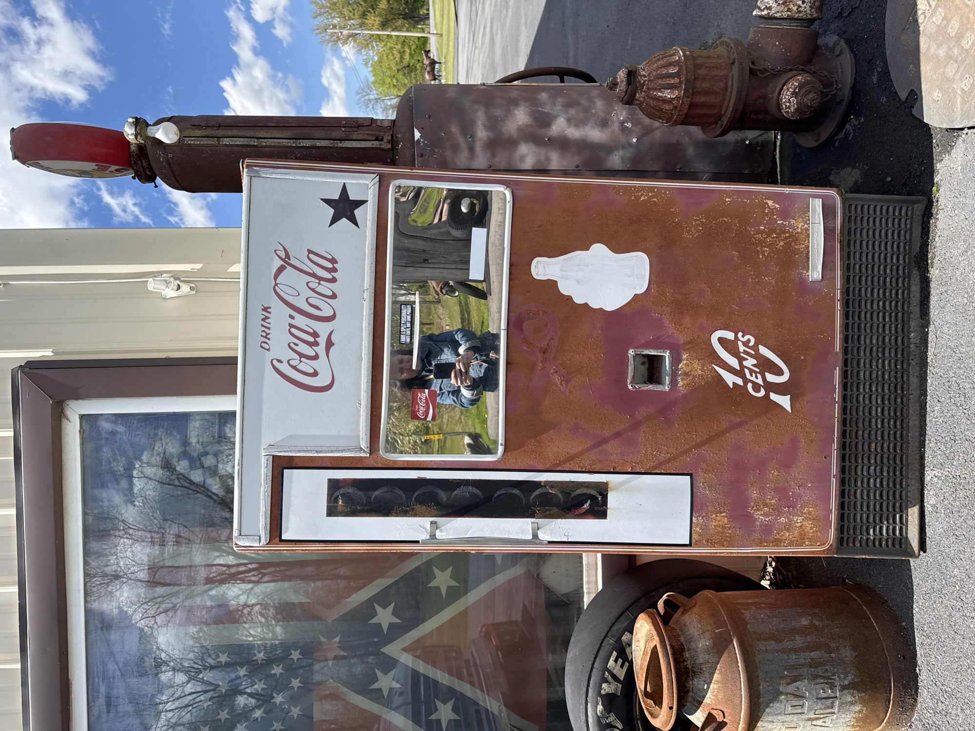 This image shows a vintage Coca-Cola vending machine with rust damage, suggesting it's an antique item. The machine is decorated with the Coca-Cola logo, a star, a hand-holding-bottle sticker, and the price of "10 cents." A reflection in the machine's mirror shows a person holding a phone, taking a photo of the scene. The background reveals a window reflection of a Confederate flag. An old gas pump with a red top and a rusty fire hydrant are positioned behind the vending machine. A Goodyear tire and a weathered metal milk can are situated at the base of the vending machine. The photo was taken in the Town of Seneca Falls, United States of America.