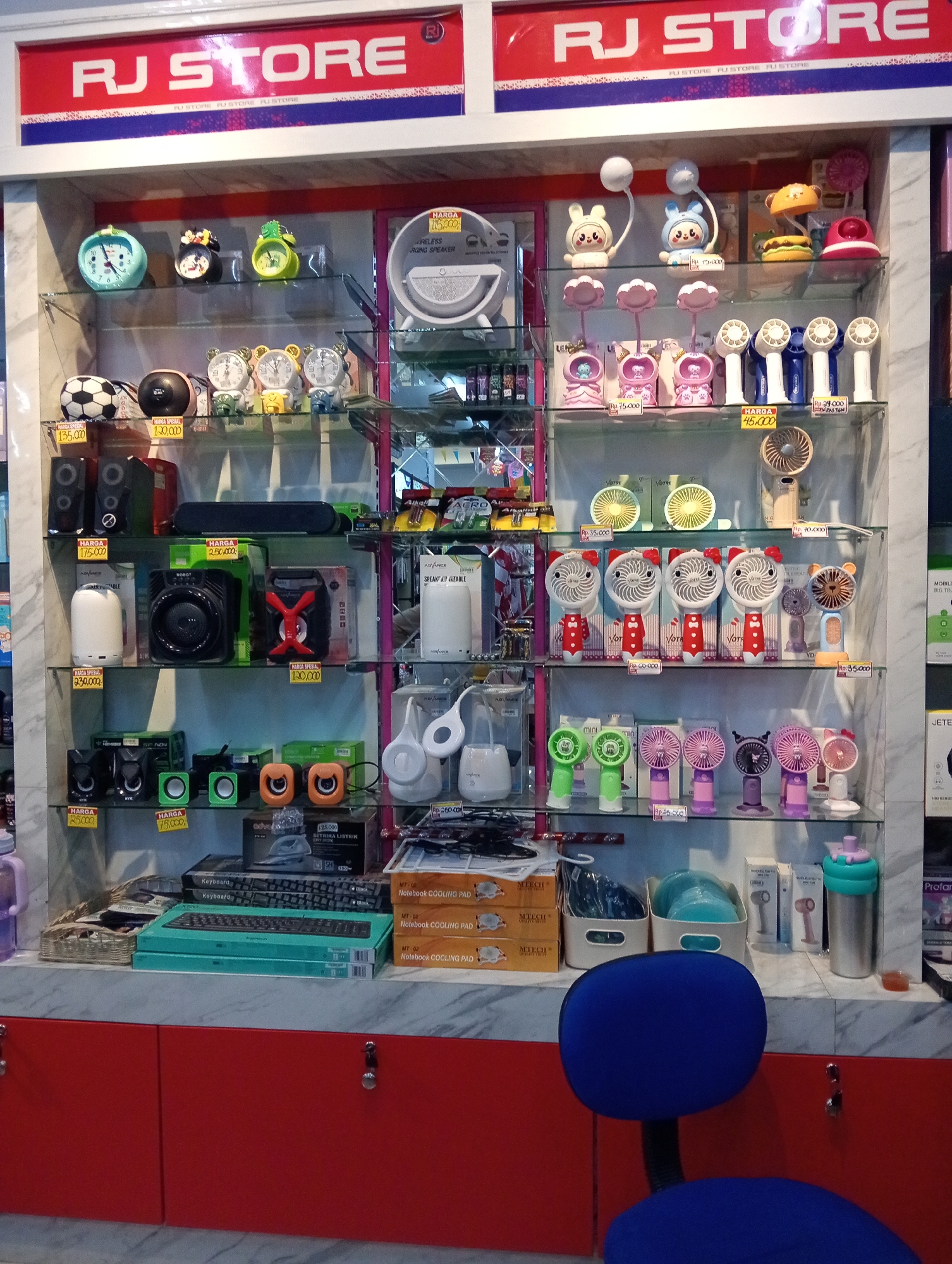 The image shows a retail store with a prominent "RJ STORE" sign at the top. The store appears to be located in Pringsewu, Indonesia, based on the provided context. The store interior is filled with shelves displaying various electronic gadgets and accessories.
The shelves are organized with distinct product categories. The top shelf features decorative alarm clocks in cartoonish designs, including Mickey Mouse and teddy bear characters, alongside a portable speaker. The second shelf displays a collection of small, handheld fans with cartoonish designs, as well as some larger USB fans and desk fans. These fans come in various colors, including white, blue, and beige, and some have price tags indicating they are for sale.
The third shelf is stocked with different types of speakers, including small, colorful computer speakers and larger, black portable speakers. There are also some smaller electronic devices and accessories on this shelf. The fourth shelf holds a variety of smaller fans, some designed to be held in the hand, in pastel colors like pink, purple, and green, along with some black and white models.
Further down, the shelves contain keyboards, notebook cooling pads, and other electronic accessories. One shelf also has a basket filled with what appear to be small bags or pouches. There are also some larger appliances like an iron visible on a lower shelf.
The overall environment is brightly lit, suggesting it is a well-maintained retail space. The store is presented as a place offering a wide range of small electronic goods.