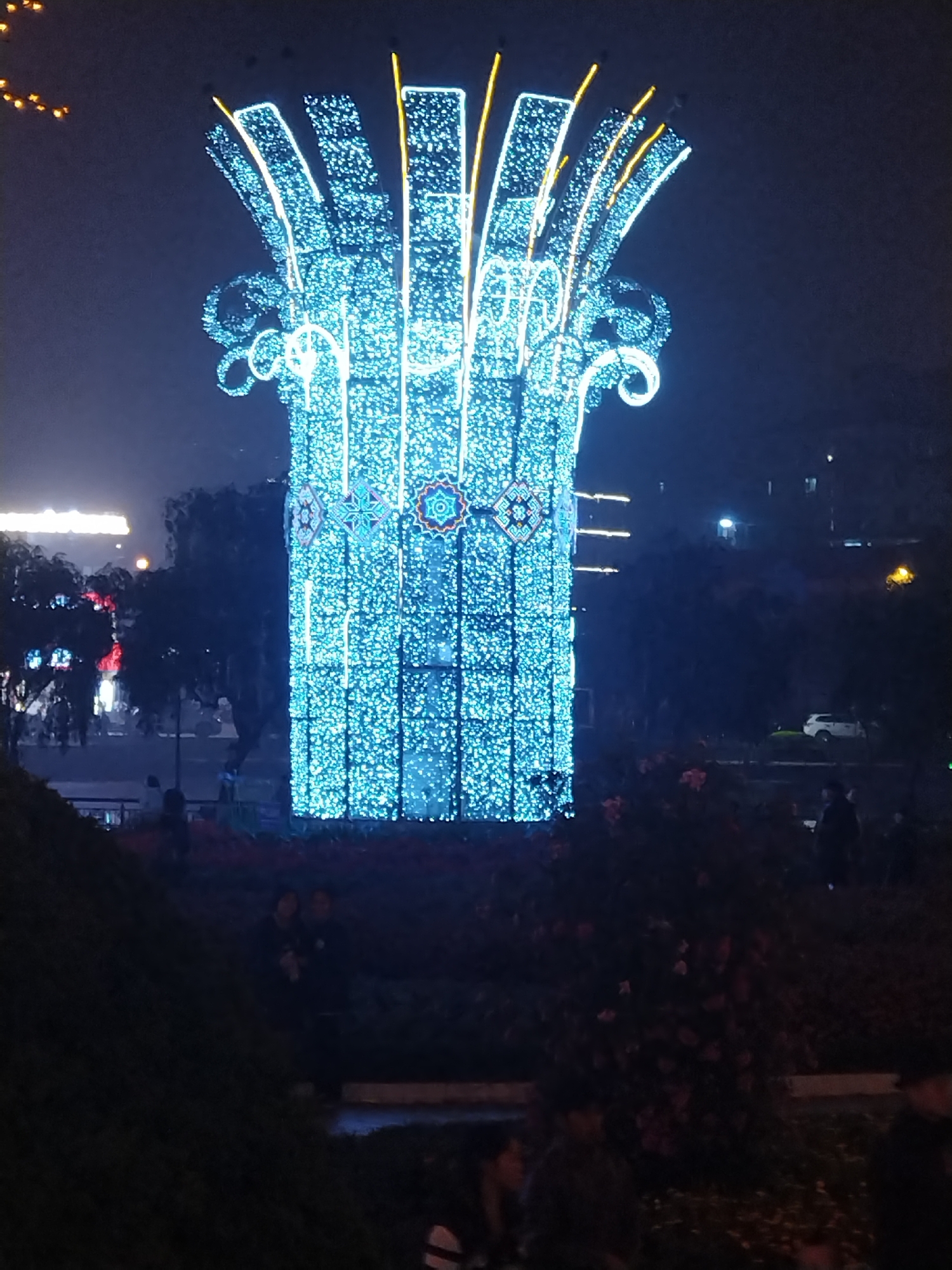 At night in Sa Pa Ward, Vietnam, a tall, light-blue light display is present. The display has a cylindrical base covered in small lights, with decorative patterns near the top. The top of the display features curving arms with more lights and some yellow rods extending upward. People are visible in the background and in the foreground, partially obscured by dark shrubbery. A building is partially visible in the background behind the light display. A white car is parked on the right side of the scene in the background.