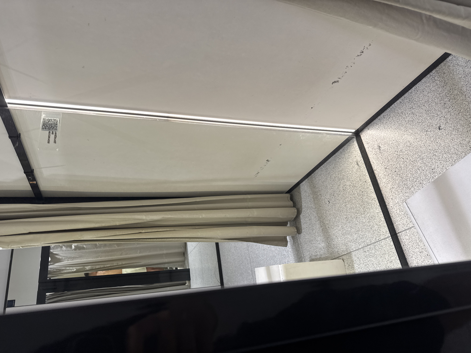 The image captures an interior space from a low vantage point, looking upwards. The ceiling is white, featuring a long, linear LED strip light. Visible on the white ceiling are several dark scuff marks or dirt smudges.
In the upper left, a QR code is present on a transparent panel, likely glass or plastic, positioned near the ceiling light. Adjacent to the QR code, upside-down Cyrillic text reads "ВОЗМОЖНОСТИ ДОБАВЛЕНЫ," which translates to "POSSIBILITIES ADDED."
Below the ceiling, beige or light tan curtains are drawn across, partially revealing a person's lower legs (calves and ankles) in light-colored clothing behind them, suggesting someone is using the private space. The walls of the cubicle or room are light grey with a speckled, textured finish. The floor, also light grey and speckled, appears to be made of terrazzo or a similar material. Dark frames or borders outline the structural elements of the space. A white, rectangular object is situated on the floor near the curtains.
The scene depicts what appears to be a changing room, fitting room, or a private cubicle, possibly in a public or retail setting in Plovdiv, Bulgaria. The immediate foreground is a blurry, dark surface.