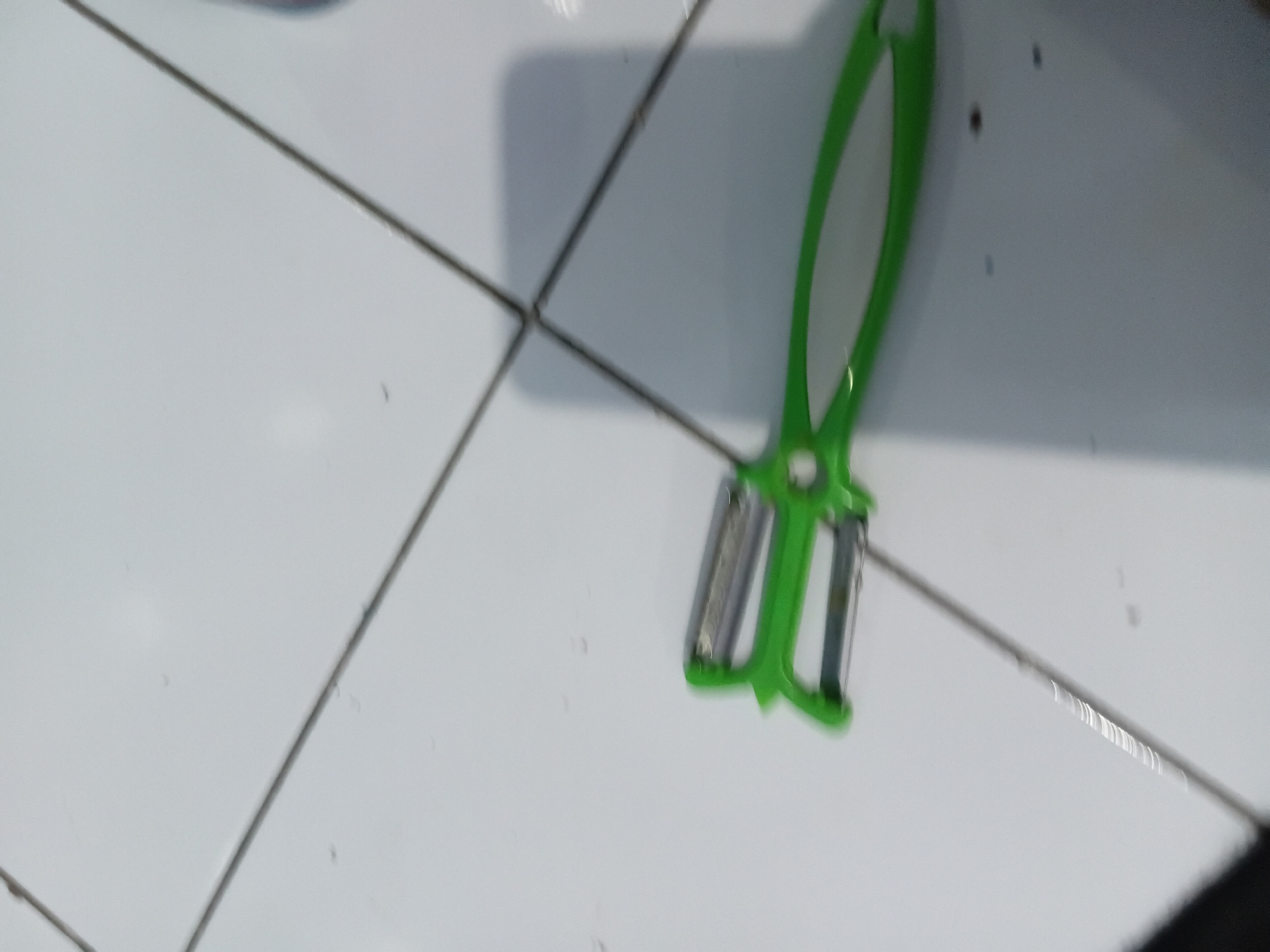 A green and silver vegetable peeler lies on a white tiled floor, which appears to be the floor of a kitchen or bathroom in a home in Tanggul Wetan, Indonesia. The tiles are laid in a grid pattern with thin, dark grout lines. The peeler is positioned diagonally across the frame, with the silver blades facing upwards and the green handle curving towards the bottom right. The lighting suggests an indoor environment, possibly with sunlight casting a shadow from an unseen object to the right of the peeler. The overall scene is simple and domestic, depicting a common kitchen utensil in an everyday setting.