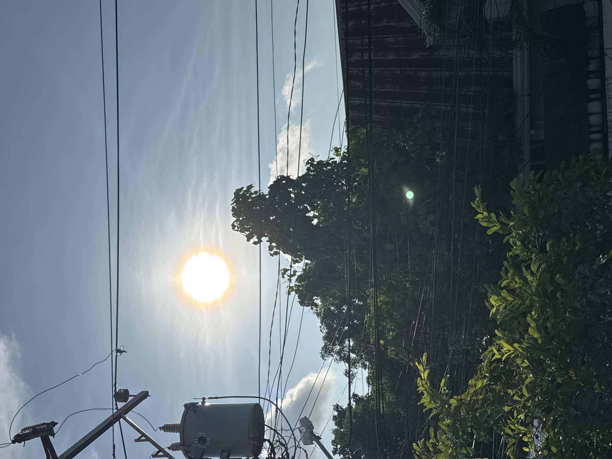 The image depicts an urban scene in Davao City, Philippines, characterized by a bright, obscured sun positioned high in a partly cloudy, blue sky. Power lines crisscross the view, running from the lower left to the upper center. A utility pole and cylindrical transformer are located in the lower left corner. Dense foliage and trees dominate the right side of the frame, partially obscuring a building with a dark facade. The sun's glare creates a halo effect, while a secondary green reflection is visible in the upper right portion of the image, possibly reflecting off foliage or glass.