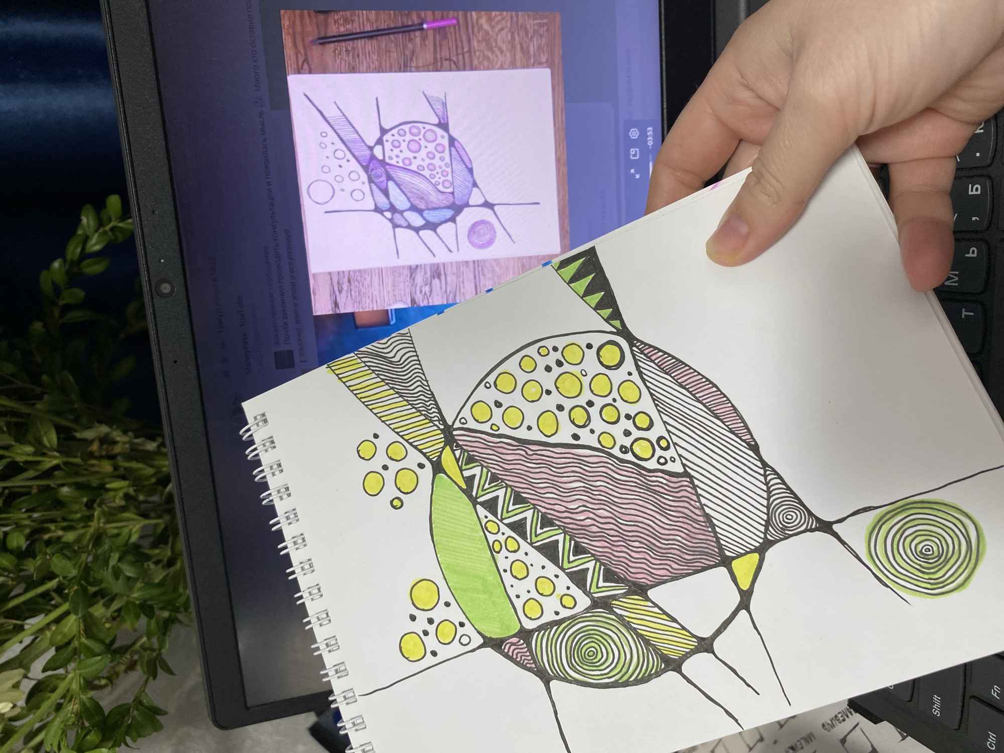 An overhead shot captures a hand holding a spiral-bound notebook with an abstract drawing, placed in front of a laptop. The drawing in the notebook features black outlines forming organic shapes, filled with various patterns including stripes, circles, waves, and zigzags, colored in yellow, green, and pink/reddish hues.
The laptop screen displays a YouTube video featuring a similar abstract drawing, predominantly in purple and light blue tones. Video controls are visible, showing a timestamp of "-03:53". Text snippets from the laptop screen include a user or channel name "MoneyPass - YouTube" and messages in Russian: "Тимур вчера в 18:28" (Timur yesterday at 18:28), "Закрепленное сообщение" (Pinned message), "Почти закончил проводить консультации и появилась мысль" (Almost finished conducting consultations and a thought appeared), and partially visible "Много кто оставил пов" (Many people left pov) and "1 тысячи, вот в этом и вся разница" (1 thousand, that's the whole difference).
A light-skinned hand holds the top right corner of the notebook. Part of the laptop's keyboard is visible to the right, showing Cyrillic characters (б, ь, M, T) and English keys (Shift, Ctrl, Fn). The brand "SAMSUNG" and text "MALEVIK" are visible on the keyboard area. To the left, a blurred green leafy plant adds a touch of natural element to the indoor setting. The scene suggests a creative activity, possibly following an online art tutorial or comparing an artwork to a digital reference in Noviy, Russia.