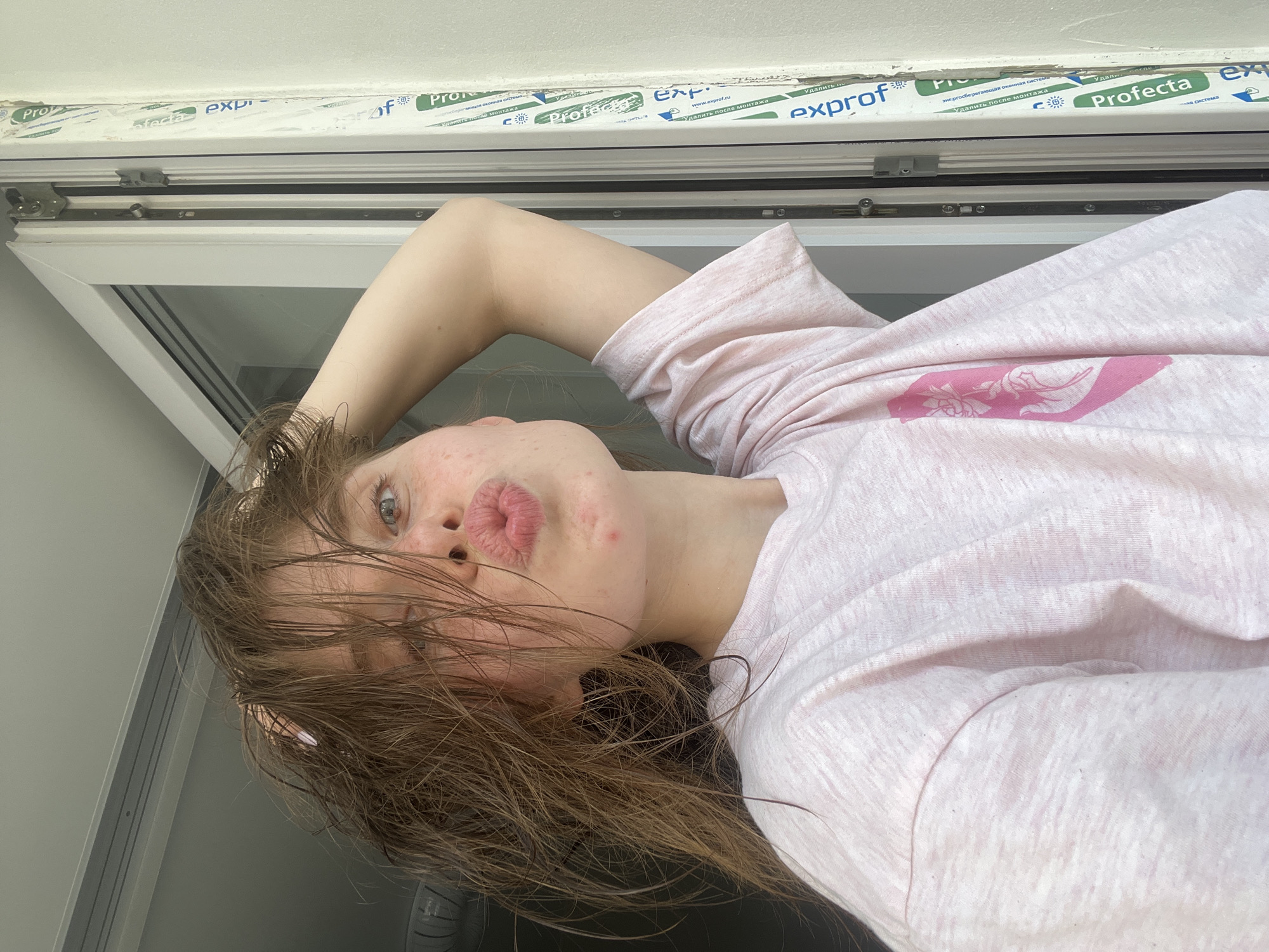 This is a close-up, vertically oriented selfie of a young person, likely female, with long, messy brown hair. The person is wearing a light pink t-shirt with a pink graphic design on it. Their face is turned towards the camera, with their eyes open and looking at the viewer. They are puckering their lips in a kiss pose. The skin on their face shows some blemishes and redness. Their right arm is raised and bent, with their hand resting on their head, pushing their hair back.
The background appears to be a window frame and the sky. The window frame has visible text on a protective film, which includes "Prof, "Profecta", and "exprof.ru", along with Russian text that translates to "remove after installation". This suggests the photo was taken shortly after window installation. The overall lighting indicates it is daytime, likely outdoors or near a window with natural light. The scene is casual and personal, likely taken in a home setting in Chaikovsky, Russia, given the context. The emotion conveyed is playful and flirty due to the kiss pose and direct gaze.