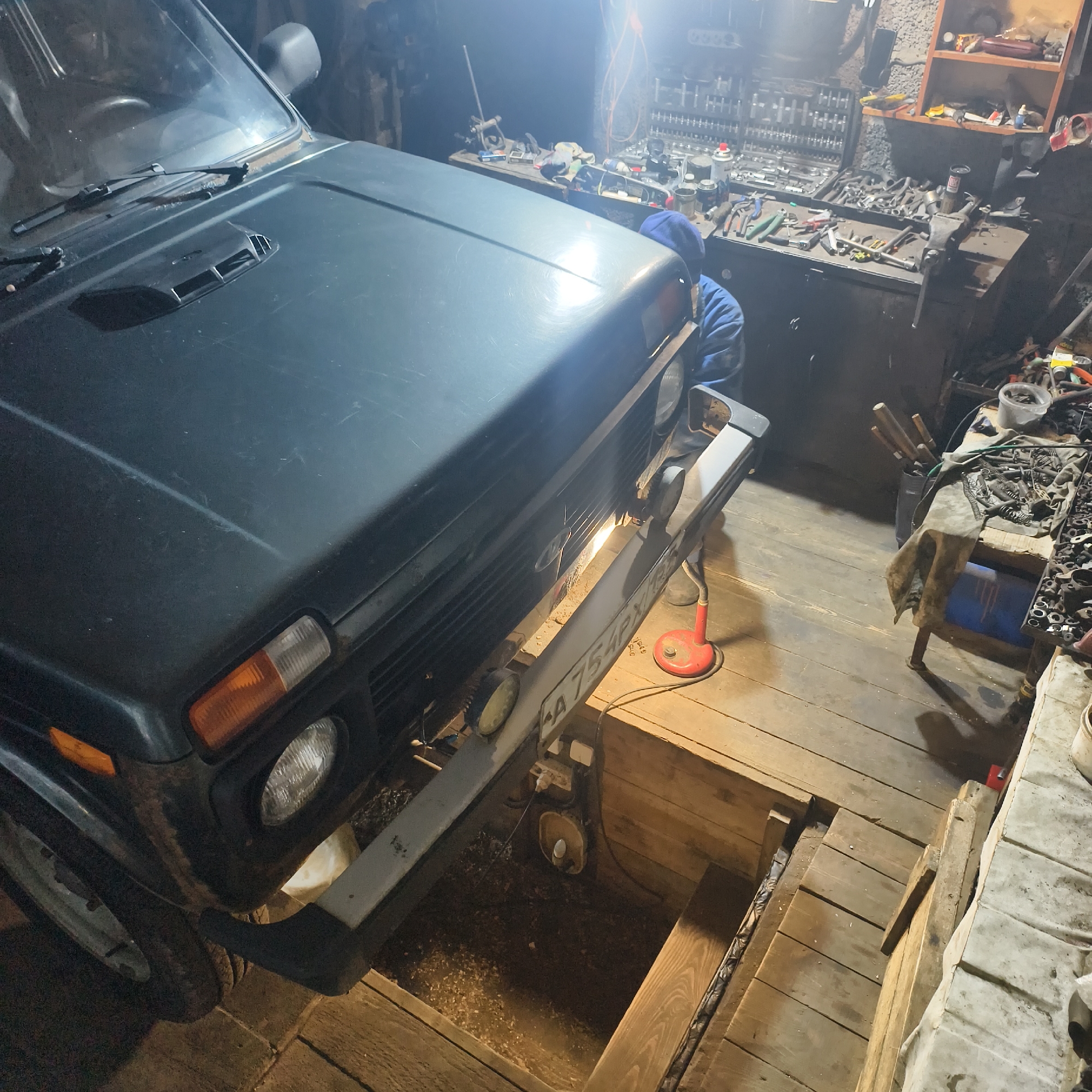 This image depicts the interior of a cluttered garage or workshop in Фоки, Russia. A dark-colored, possibly dark green, Lada Niva (indicated by the body style and grille logo) is the primary subject, positioned with its front end extending over an open, wooden-lined inspection pit. A partial Russian license plate, "A 754PX", is visible on the front bumper. A red portable work light is situated on the wooden floor, illuminating the underside of the vehicle's front bumper. In the background, to the right of the vehicle, a person wearing a blue hooded jacket is partially visible, appearing to be working at a workbench. The workshop contains multiple workbenches heavily laden with various tools, including socket sets, wrenches, pliers, and other automotive equipment. A large vise is mounted on one of the workbenches. Shelves above the work area hold additional items. The floor consists of wooden planks, and wood shavings are visible at the bottom of the inspection pit. The scene is illuminated by artificial overhead lighting and the portable work light. The vehicle's presence over the pit suggests it is undergoing inspection or maintenance.