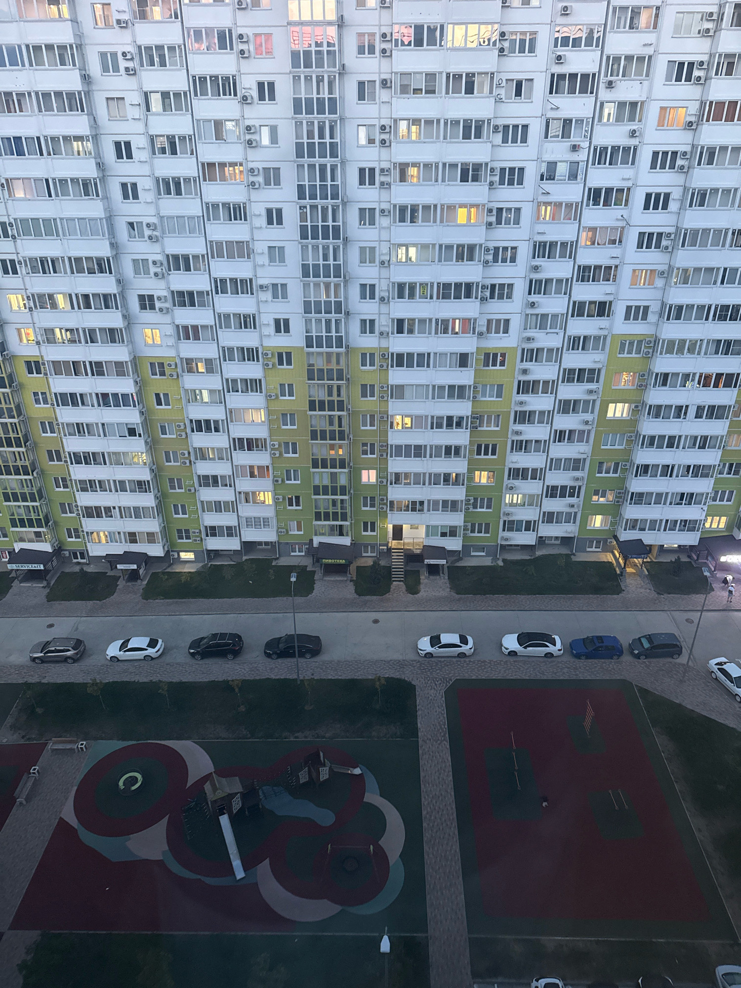 The image shows a high-angle view of a modern, multi-story residential building with a green and white facade. Numerous windows and balconies are visible on the building's exterior, with some windows illuminated. In front of the building, a paved road is lined with several parked cars of various colors and models. Further in the foreground, a children's playground with colorful ground markings and play structures is present. The overall scene appears to be an urban residential area in Anapa, Russia, likely captured during the late afternoon or early evening.