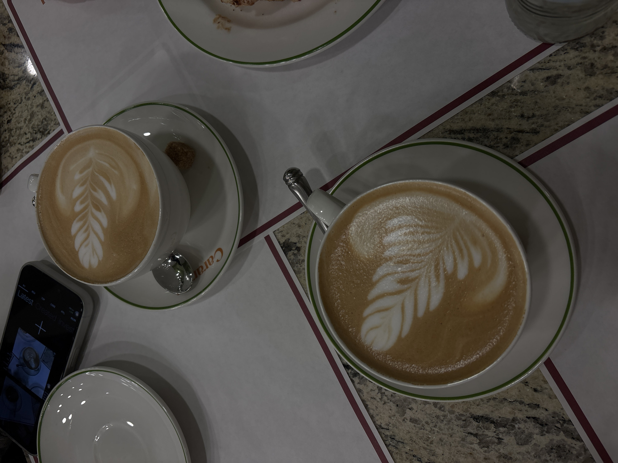 The image shows two cups of coffee with latte art on a table, likely in a cafe. The table has a white surface with burgundy and gray stripes. Next to one of the cups is a smartphone. A saucer, a spoon, and a small sugar cube are also visible near the cups. The setting appears to be indoors, with granite table. The saucer has "Cara" on it, suggesting the coffee could be at Caras, a cafe in Yerevan, Armenia.