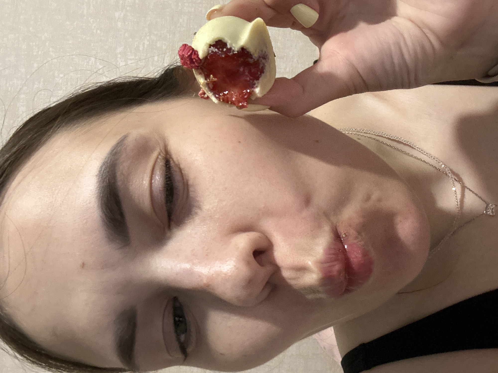 A close-up, eye-level shot shows a young woman with dark hair, her face angled towards the camera. She is wearing a black tank top and a silver necklace. Her left eye is partially closed, and her lips are puckered as if about to kiss or taste something. In her right hand, she holds a white chocolate truffle with a raspberry topping, a bite taken out of it, revealing a red filling. Her fingernails are painted yellow. The background is a plain, textured wall. There is no discernible indication of the city of Perm, Russia, in the image.