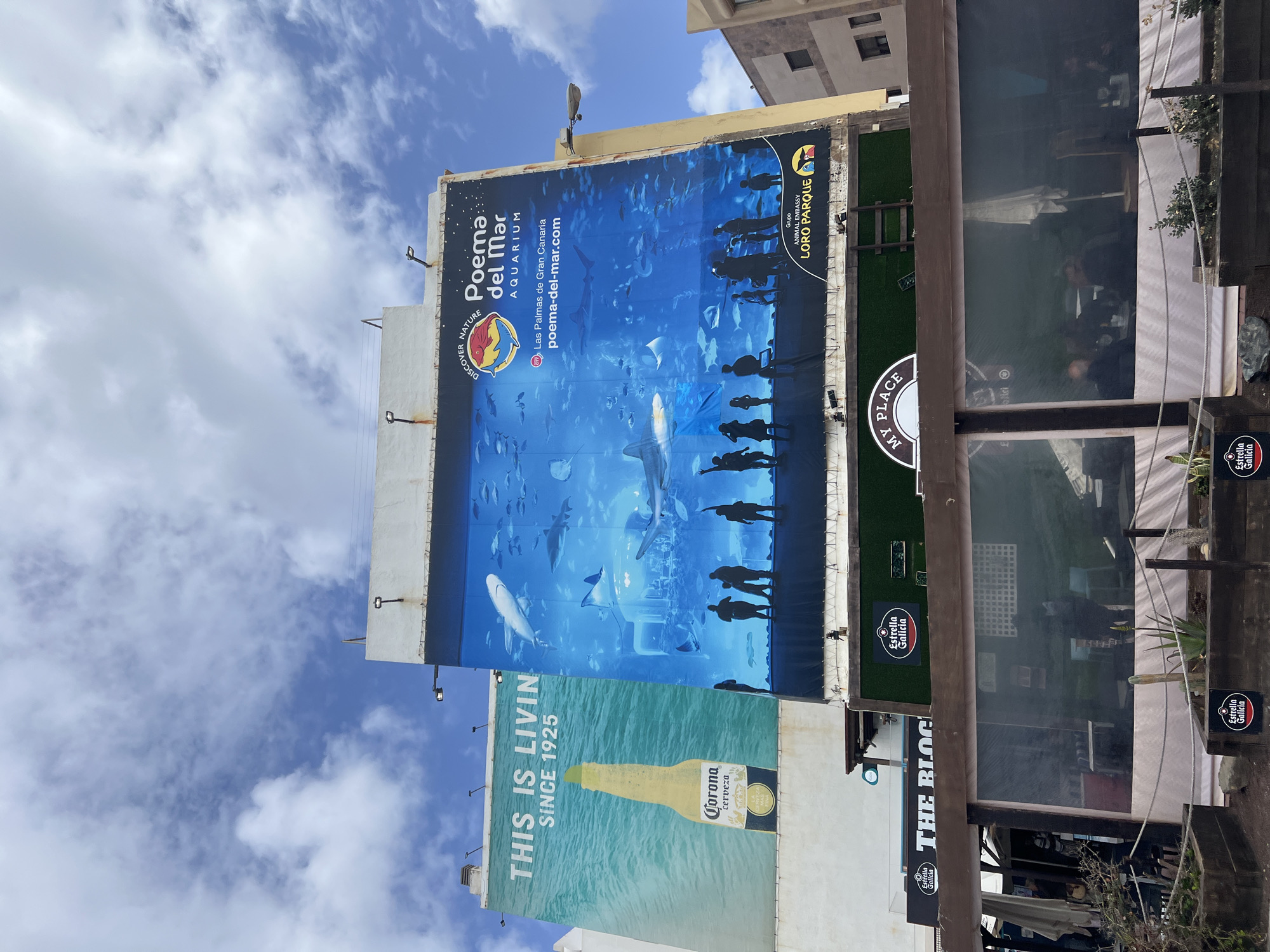 The image shows the exterior of a bar or restaurant in Las Palmas De Gran Canaria, Spain. Several advertisements adorn the outside of the building. The top ad is for Poema del Mar Aquarium. Below that is an advertisement for Corona beer. The restaurant is called "The Bloc". There is a sign for "My Place". The image also shows Estrella Galicia beer advertisement.