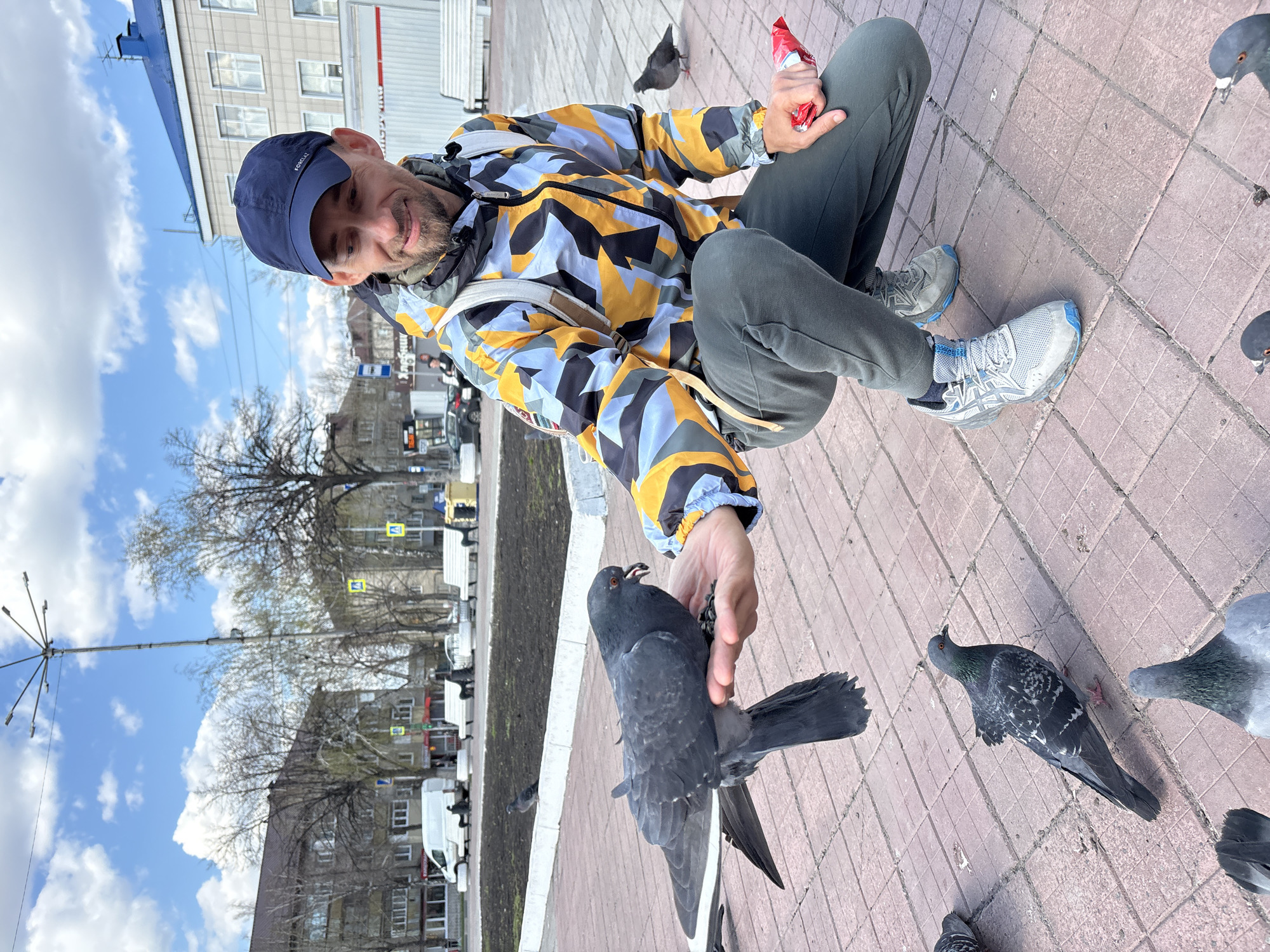 The image depicts a smiling man feeding pigeons in an urban setting in Ulyanovsk, Russia, on a clear, sunny day. The man, who appears to be in his 30s or 40s with a beard, is squatting on a reddish-brown tiled pavement. He wears a dark blue baseball cap with "FORCLAZ" visible on it, a colorful geometric patterned jacket (yellow, black, blue, and grey), dark grey pants, and light grey and blue athletic shoes.
In his left hand, the man holds a crumpled red packet, possibly containing food. A dark grey pigeon is perched on his outstretched right hand, actively eating from it, while other pigeons are scattered on the ground around his feet. The man's cheerful expression conveys a sense of enjoyment from the interaction.
The background reveals a typical city landscape with multi-story buildings, some with visible windows, and several bare trees, suggesting late winter or early spring. A tall, multi-headed lamp post stands against the bright blue sky, which has scattered white clouds. Further in the distance, a street is visible with vehicles, including white vans, and road signs, one featuring a black arrow pointing right. The overall scene captures a pleasant moment of human-animal interaction within a bustling urban environment.