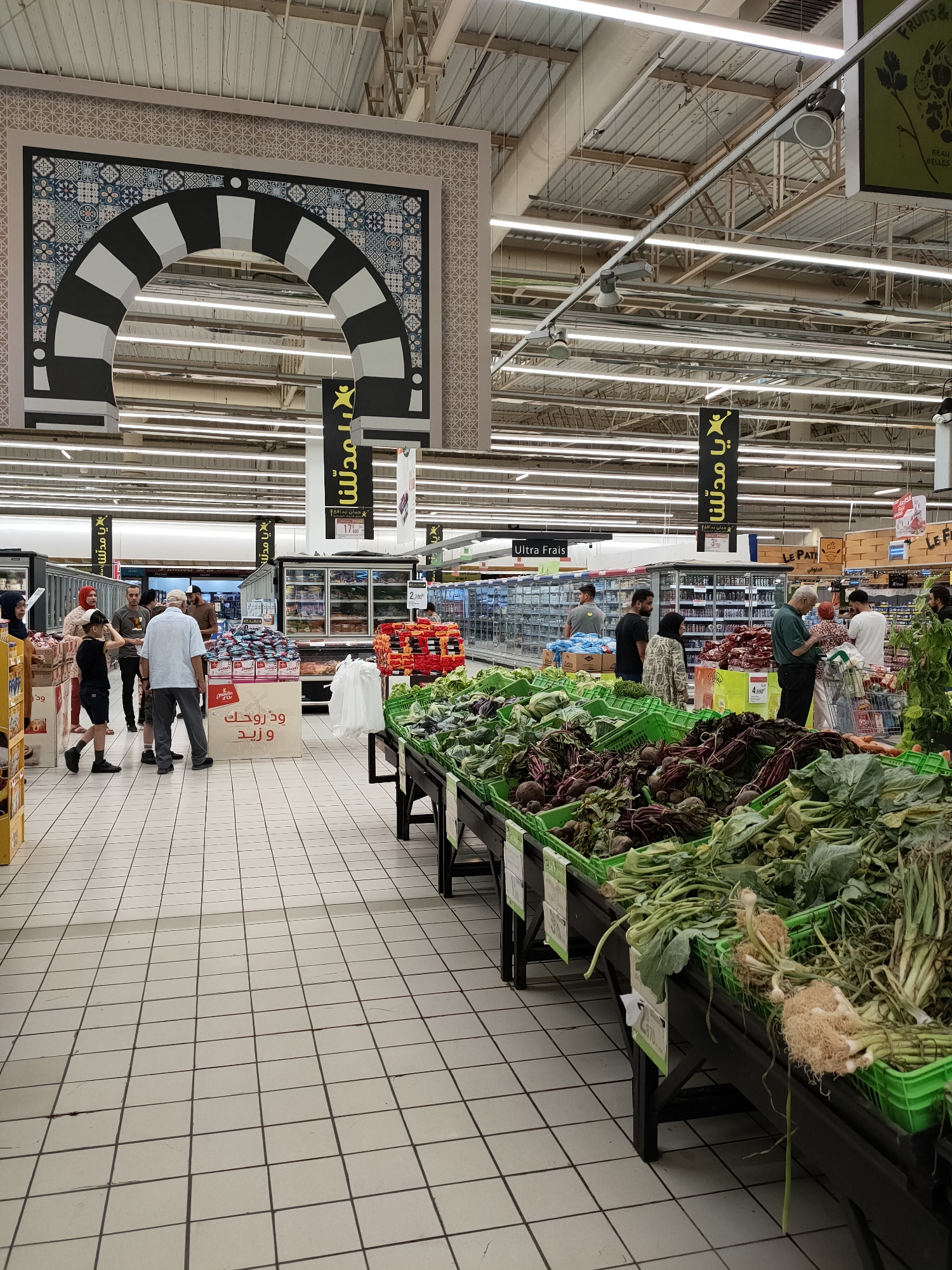 The image depicts the interior of a supermarket in Cebalat, Tunisia. The scene is dominated by aisles filled with produce and shoppers. In the foreground, a wide variety of fresh vegetables are displayed in green crates on dark wooden stands. These vegetables include beets, leafy greens, and root vegetables with their tops still attached. Shoppers, dressed in casual attire, are seen browsing the produce section and interacting with each other.
Further back in the supermarket, aisles are lined with refrigerated display cases stocked with food items. Several banners with Arabic script hang from the ceiling, indicating different sections or promotions. An ornate archway with a black and white striped pattern and intricate blue and white tiling is visible on the left side of the image, suggesting a themed or decorative element within the store. The lighting appears to be fluorescent, typical of a commercial environment. The floor is tiled with a light-colored grid pattern. The overall impression is of a bustling marketplace within a retail setting.