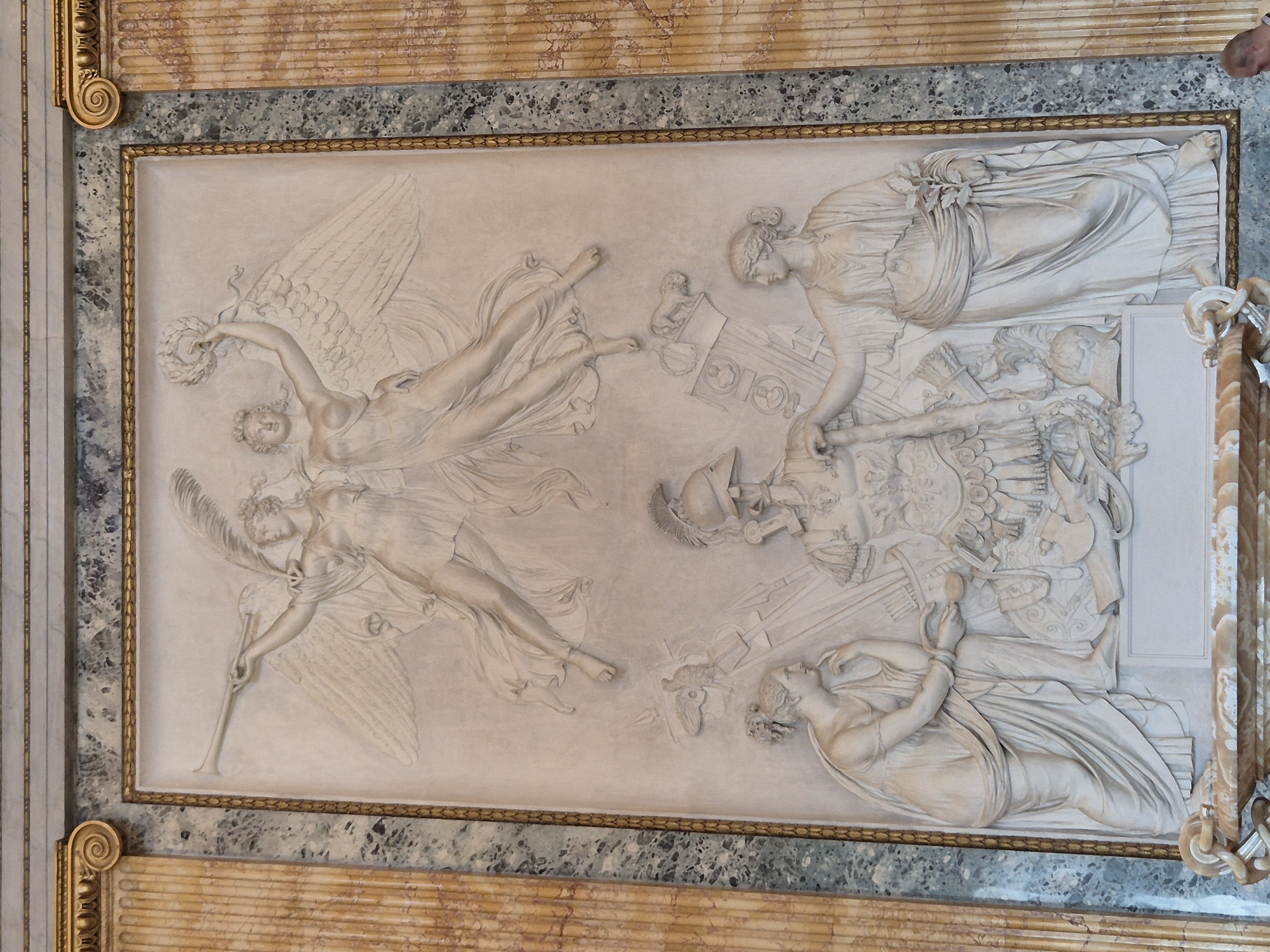 This is a photograph of a detailed bas-relief sculpture adorning a ceiling, likely within a grand interior space in Caserta, Italy. The artwork depicts a mythological or allegorical scene featuring several figures.
On the left, two winged figures, possibly nymphs or goddesses, are shown in dynamic poses. One is in flight, holding a wreath, while the other appears to be reaching out. Their drapery flows elegantly, suggesting movement and ethereal qualities. On the right side of the relief, a group of figures is arranged around a central, armored figure, likely a warrior or a deity. One figure reclines below, seemingly receiving attention or possibly in a state of defeat or slumber, holding an object, possibly a mirror or a jewel. The overall composition suggests a narrative of victory, tribute, or a celestial event.
The sculpture is rendered in a classical style, with an emphasis on musculature, idealized forms, and flowing drapery. The figures are depicted in various stages of action and repose, creating a sense of depth and drama. The material appears to be plaster or stucco, painted a uniform off-white, which highlights the intricate details of the carving and the play of light and shadow.
The relief is framed by an ornate border featuring gilded scrollwork and decorative patterns, further emphasizing its placement within an opulent architectural setting. Above and below the relief are bands of textured marble or stone, and further decorative elements indicative of a palatial or significant building. The lighting appears to be ambient interior light, suggesting the image was captured during the daytime within the building. No visible text is present in the artwork itself.