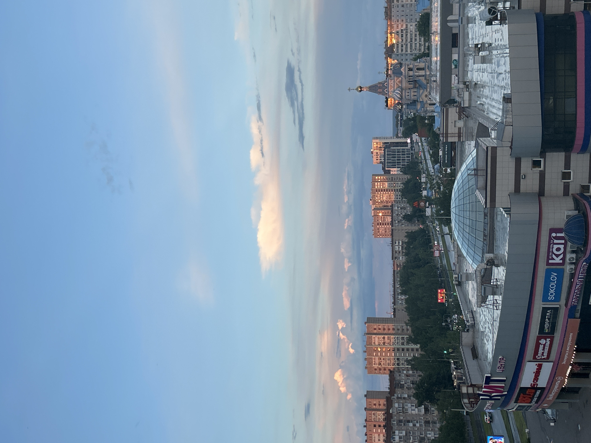 The image depicts an elevated view of a cityscape in Izhevsk, Russia, during the late afternoon or early evening, judging by the light and sky conditions. The sky is primarily blue with clouds tinged pink by the setting or rising sun. The visible urban area is comprised of multi-story apartment buildings and a significant shopping center. The shopping center occupies the foreground of the right side of the image and features various commercial signs in Russian. Notable brand names include "Sokolov," "Kari," "Kvartal," "Vse Instrumenty" (All Tools), and a large dome structure. Further away, a Russian Orthodox Church with a prominent golden dome and cross is visible amongst the buildings. A roadway can be seen near the bottom of the image, with vehicular traffic. The overall scene suggests a mix of residential and commercial areas typical of a modern urban setting.