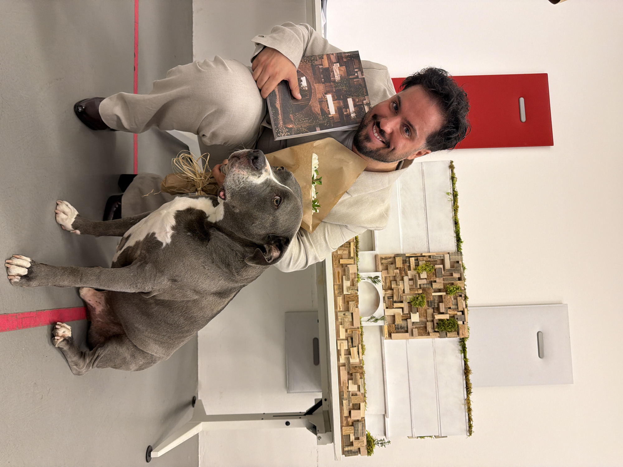 An indoor photograph captures a smiling man and a dog posing together. The man, with dark curly hair and a beard, is wearing a light beige or off-white suit over a dark shirt and brown dress shoes. He is sitting or kneeling on a grey floor with a prominent pink line running across it. He holds a book with an abstract cover featuring a pattern of varied wooden blocks and greenery, and a small bouquet of white flowers wrapped in brown paper with raffia. His gaze is directed towards the camera with a wide, friendly smile.
Beside him is a grey and white, short-haired dog, appearing to be an American Staffordshire Terrier or a similar breed. The dog is looking upwards with an alert expression. The background features light-colored walls and a modern, decorative panel made of numerous small wooden blocks interspersed with green moss or small plants. To the right, a red rectangular board with a single horizontal slot is visible on a white wall. The overall atmosphere is warm and friendly, conveying happiness between the man and his pet. No discernible text is visible in the image. The setting suggests a contemporary indoor space, possibly in Milan, Italy.