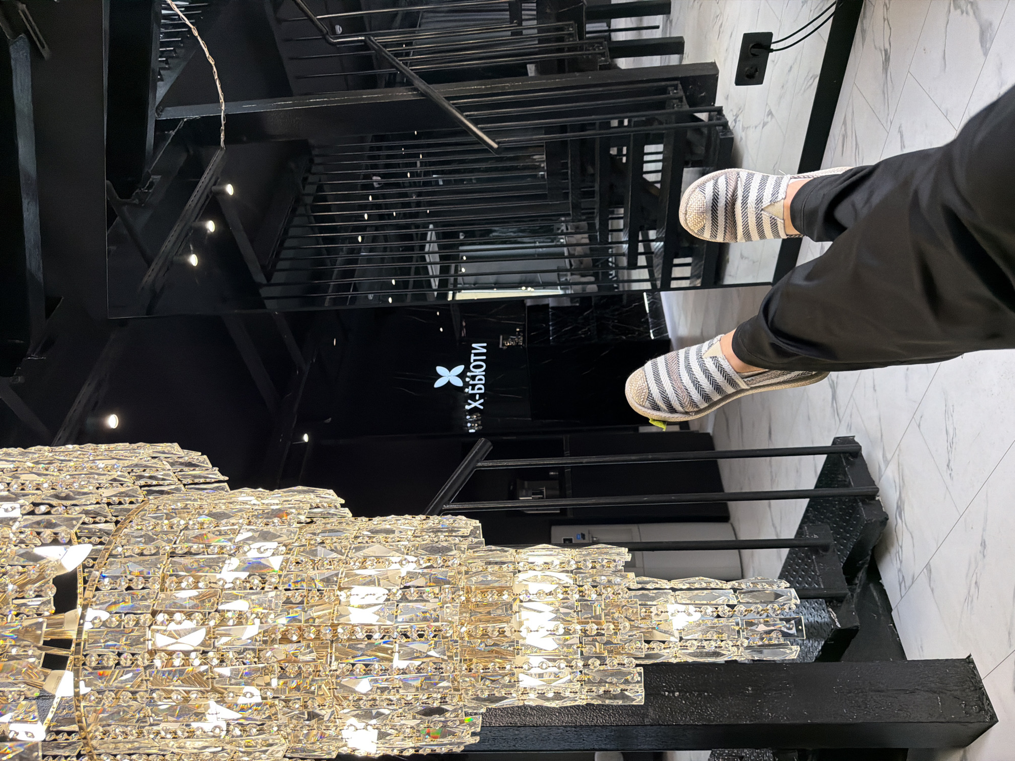 This image depicts an interior shot of a modern salon or beauty studio, likely located in Moscow, Russia. The prominent feature in the foreground is a large, multi-tiered crystal chandelier, its facets catching the light and creating a glamorous atmosphere. In the middle ground, a person's legs are visible, clad in black pants and striped slip-on shoes, suggesting they are seated or reclining, with their feet extended towards the viewer.
The background reveals a stark, contemporary design with predominantly black elements. A metal staircase with vertical bars leads upwards, its reflection visible in a large, polished black mirror wall. The logo "X-бьюти" is illuminated on the mirrored surface, confirming the establishment's identity as a beauty-related business. The floor is tiled with a light-colored marble pattern. The overall ambiance is one of sophisticated, modern luxury. The presence of a water cooler and electrical outlets suggests a functional, service-oriented space.