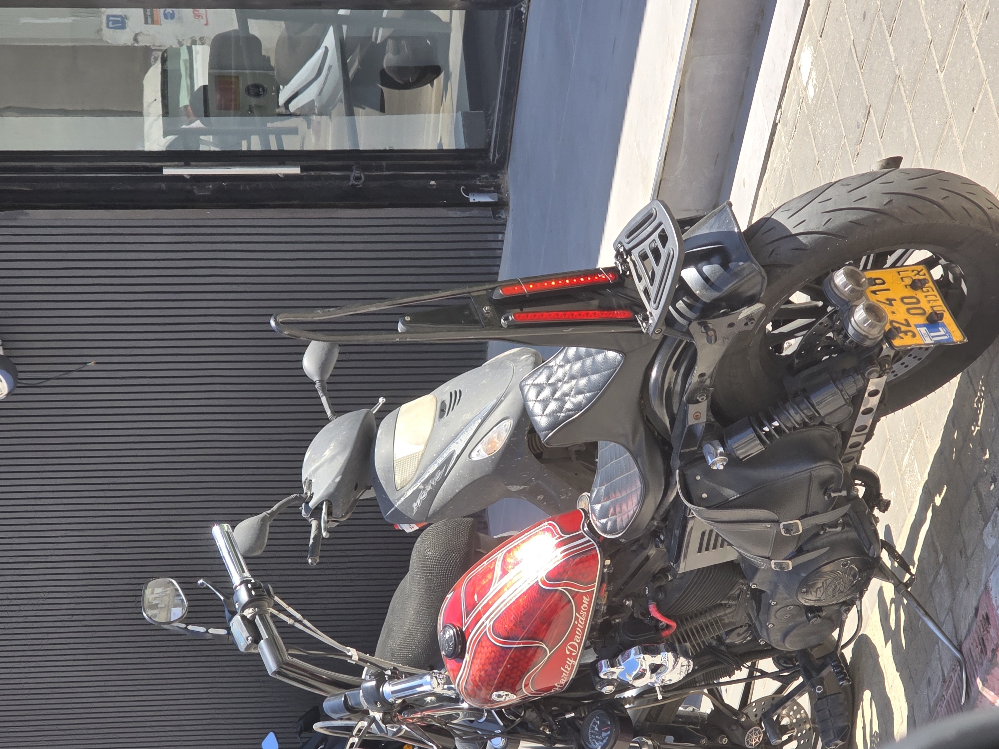 This rotated image captures two motorcycles parked side-by-side on a paved sidewalk next to a building on a bright, sunny day, likely in Tel Aviv, Israel, given the visible license plate.
The prominent subject is a custom Harley-Davidson motorcycle, positioned on the right. It features a striking red fuel tank adorned with white flame-like graphics and the text "Harley Davidson" written along its side. Chrome accents are visible on the handlebars, mirrors, and engine components. The seat is black with a distinctive quilted pattern. A black leather saddlebag, secured with buckles and possibly featuring an embossed emblem, hangs on the right side of the engine. The rear of the motorcycle showcases a thick tire, a chrome-plated kickstand, and a customized fender elimination kit with two horizontal red LED light strips serving as taillights. The yellow Israeli license plate, visible at the rear wheel, displays the number "32-416" along with Hebrew text, including "ישראל" (Israel) and an "IL" sticker, confirming the location.
To the left of the Harley-Davidson, partially obscured, is a gray or black scooter, parked very closely. Its plain design contrasts with the custom motorcycle.
The background consists of a modern building facade. The lower section features a dark, horizontally corrugated wall. Above this, a large window reflects elements of the interior, such as what appears to be furniture, signs, and possibly office equipment or merchandise, suggesting a commercial establishment. The sunlight casts sharp shadows, indicating a midday or early afternoon setting.